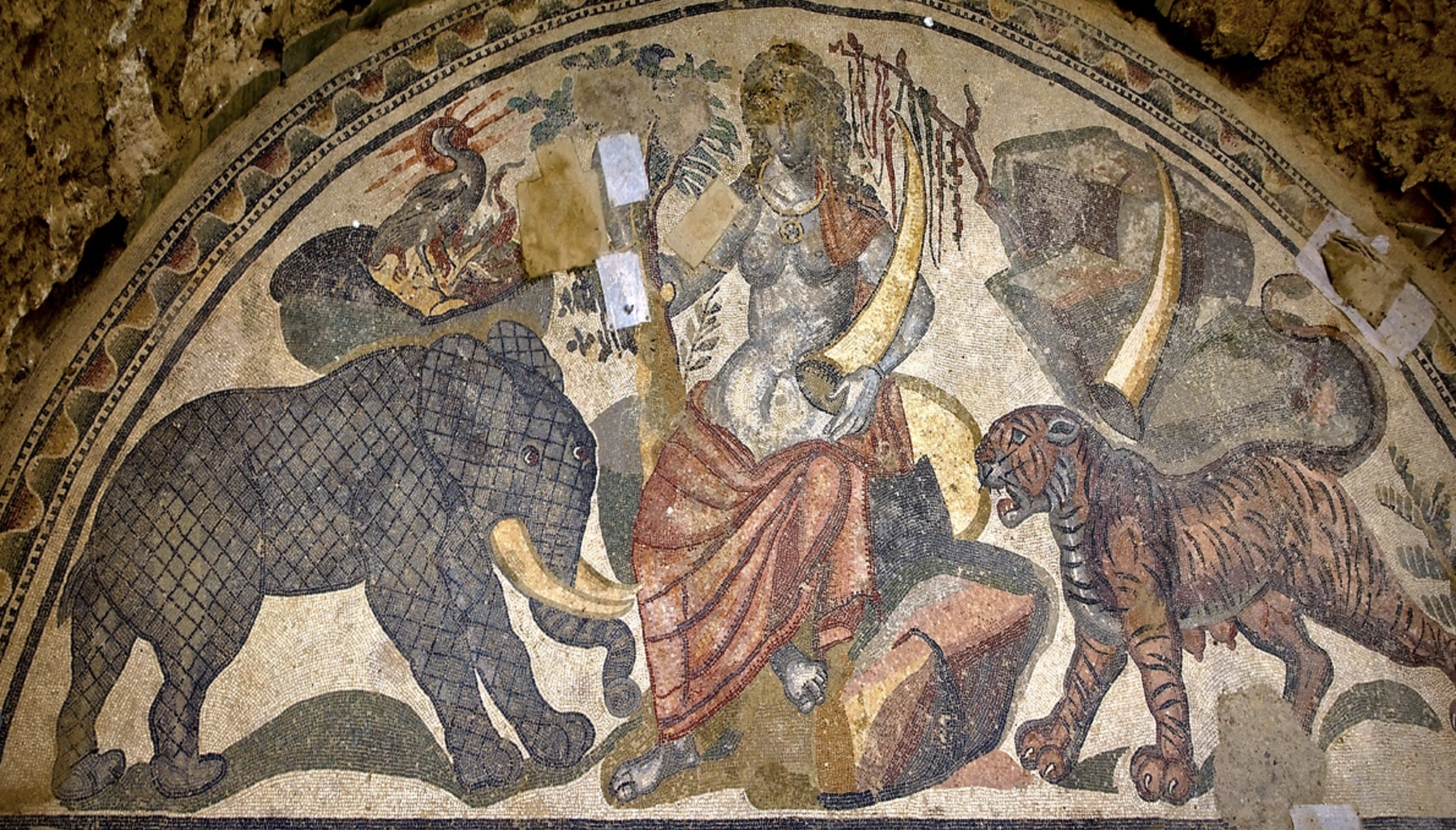 Ancient mosaic depicting a scene with a central figure surrounded by a lion, an elephant, and a peacock, all within a decorative border.