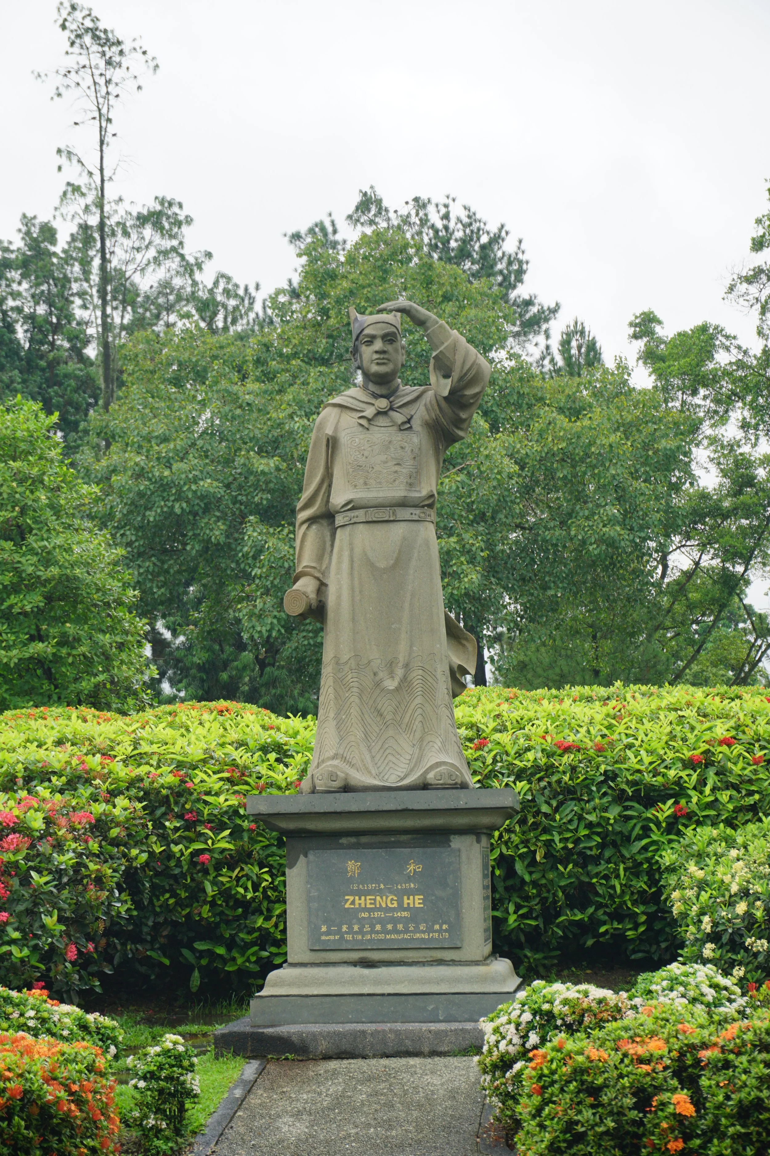 Statue of Zheng He saluting, surrounded by green bushes and trees, with a pathway in front and clouded sky in the background.