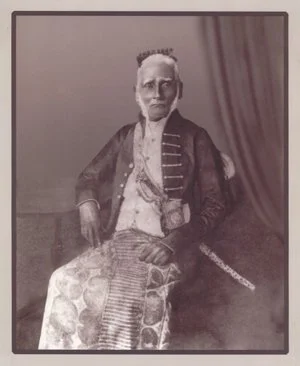 Old black and white portrait of a man in traditional clothing, sitting with a shawl draped over his shoulders and a decorated cloth on his lap.