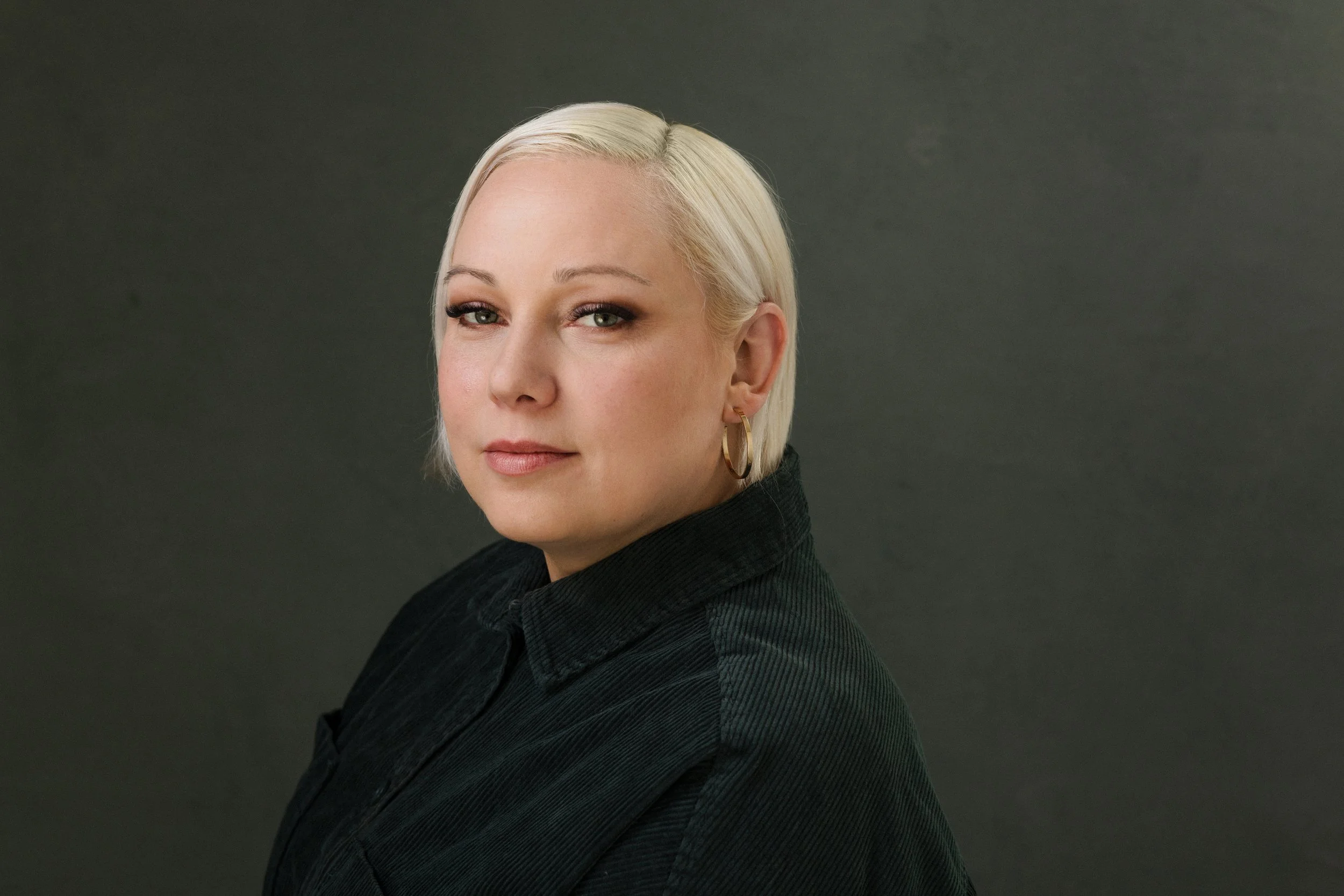 Eine junge Frau mit kurzen, platten, blond gefärbten Haaren trägt eine schwarze Jacke und große goldene Creolen, blickt leicht zur Seite, vor grauem Hintergrund.