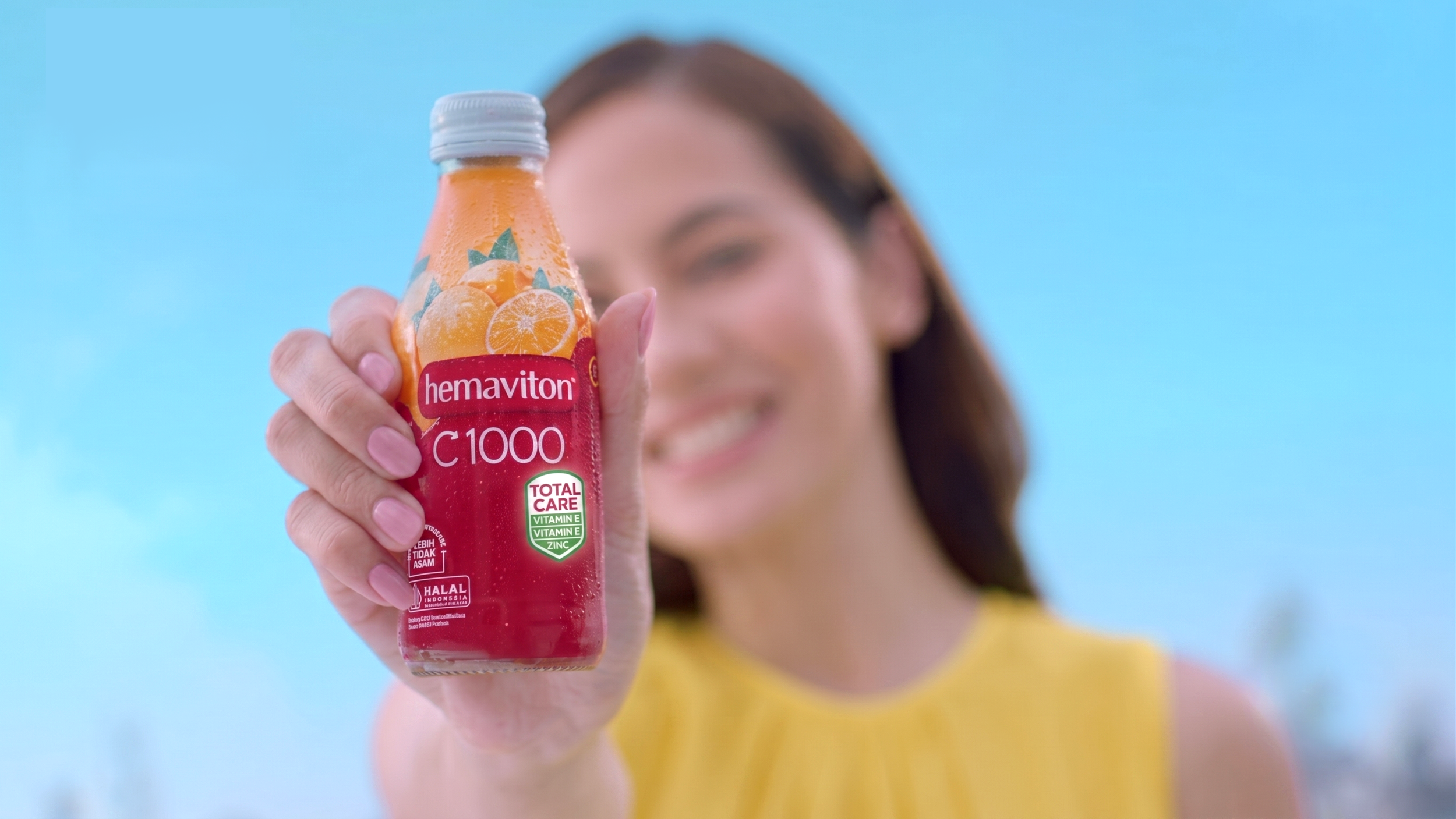 A woman holding a bottle of Hemaviton C1000 juice in front of her face, with a cheerful expression, against a blue background.
