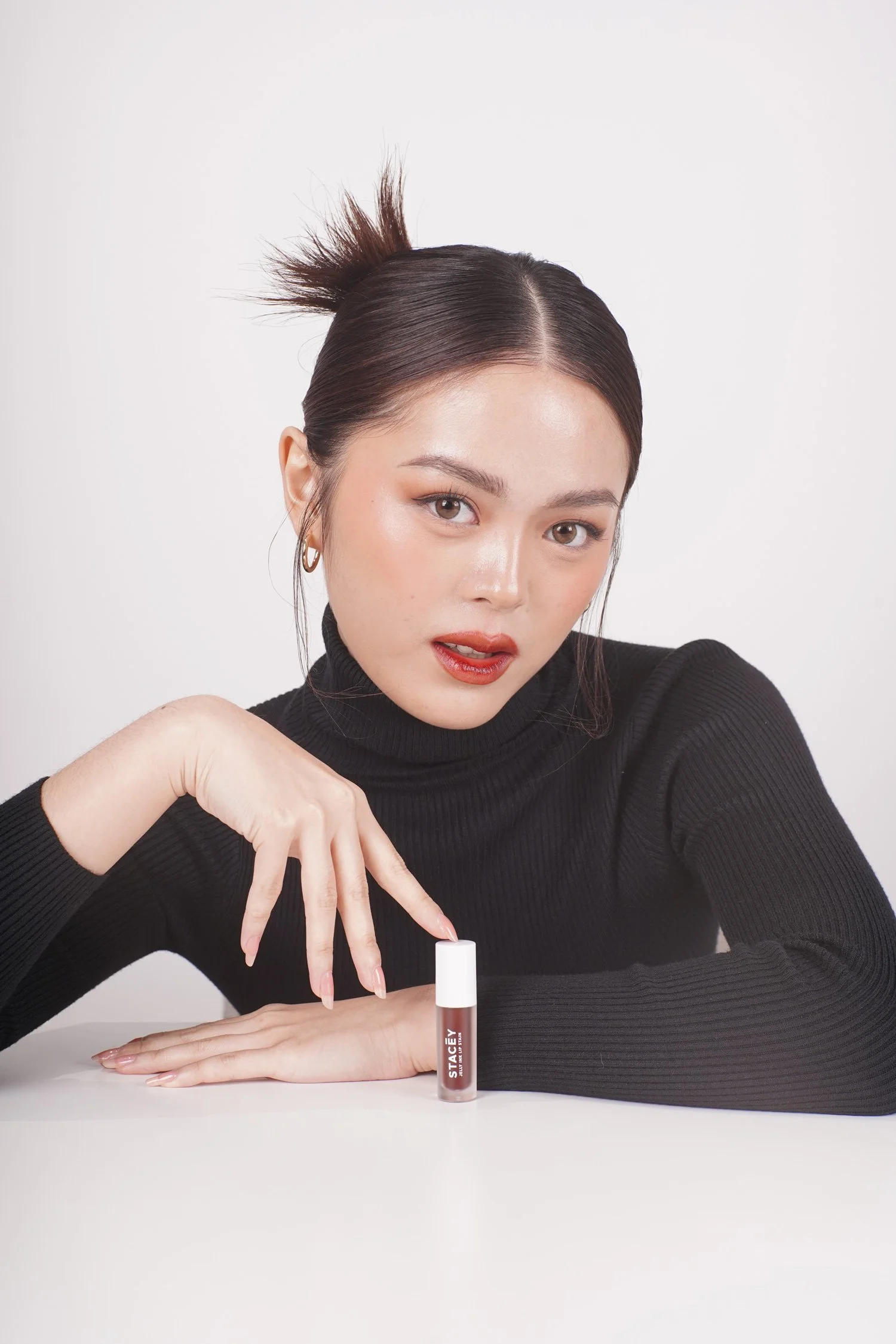 A model wearing a black turtleneck posing with a compact STACEY lip gloss bottle