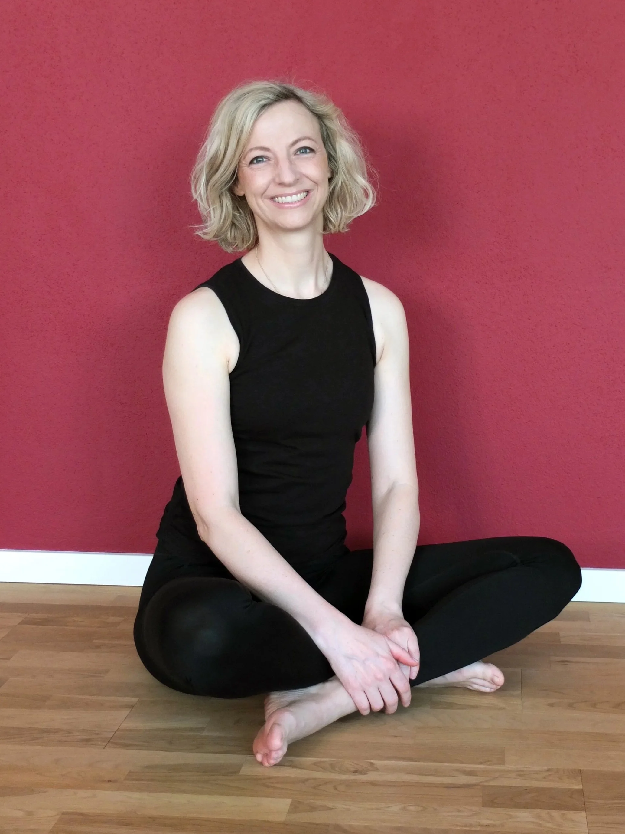 Dana Roemhild-Fries vom Pilates Loft Rosenheim sitzt entspannt auf dem Boden vor einer roten Wand mit gekreuzten Beinen und geschlossenen Händen, trägt schwarze Kleidung und lächelt freundlich.