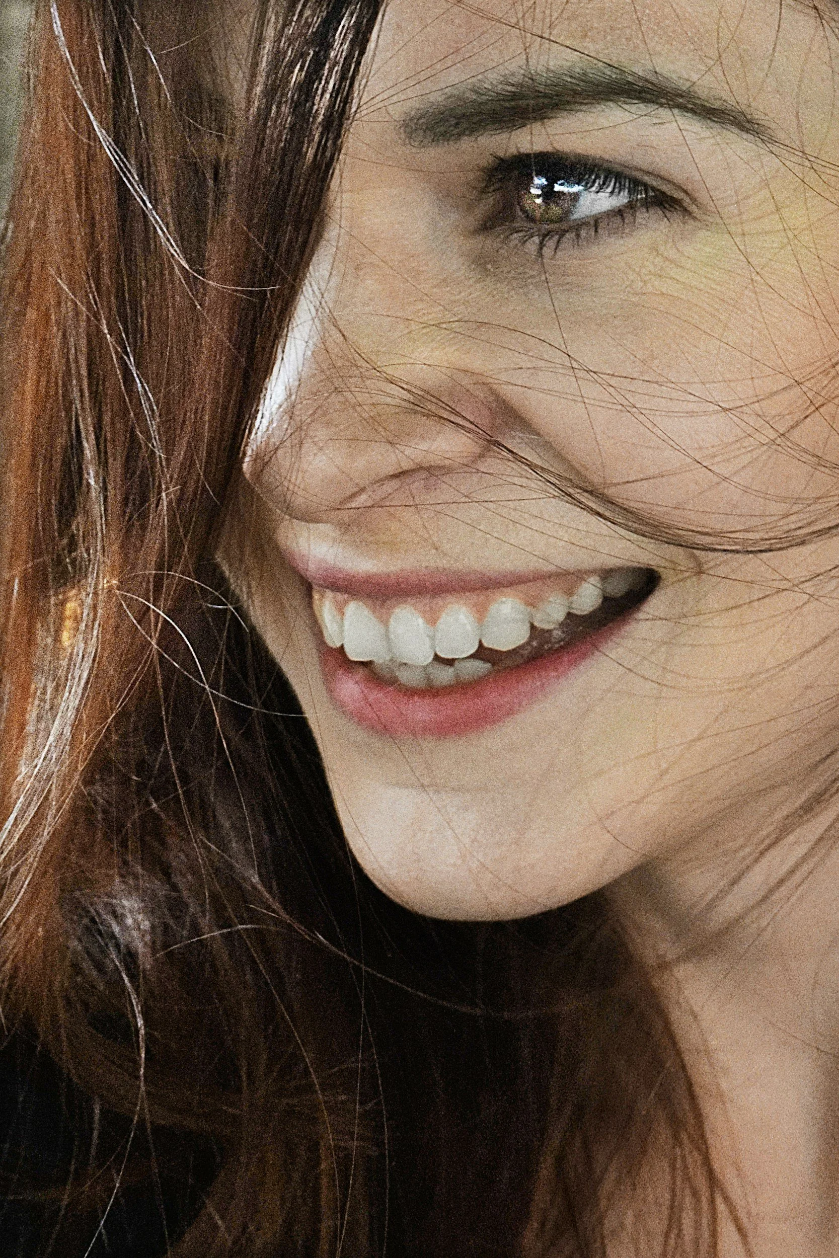 Close-up of a smiling woman with brown hair, showing her white teeth and partial facial features including eye, eyebrow, and cheek.