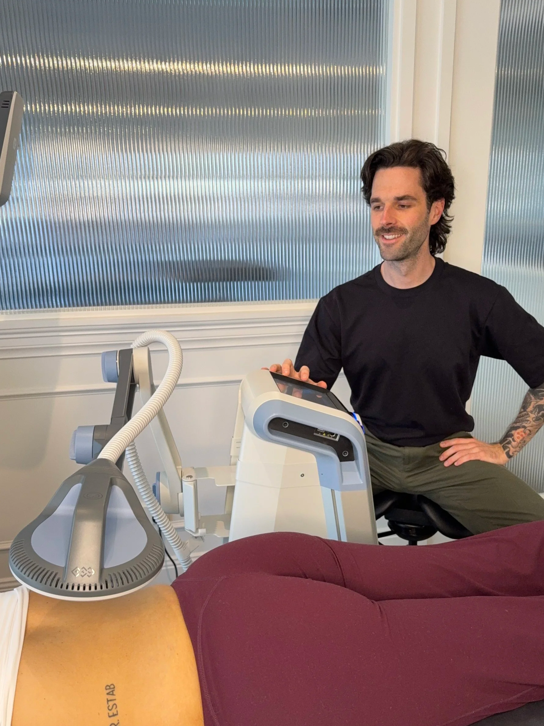 An osteopath, wearing a black shirt, using medical equipment in a clinical setting to treat a patients lower back. The person lying down is wearing maroon pants.