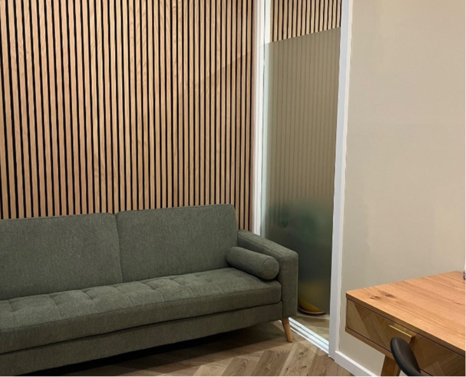 A green upholstered sofa with wooden legs, positioned against a wall with vertical wood paneling, next to a frosted glass divider, and a wooden table partially visible on the right side, in a modern interior space.