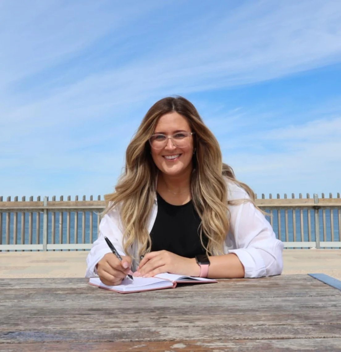 A woman with long wavy blonde hair and glasses sits at a wooden table outdoors, smiling and writing in a notebook against a background of blue sky and a body of water with a fence.