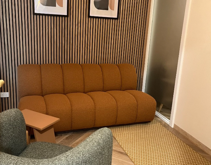 A brown sofa against a wooden slat accent wall with framed artwork, a small wooden side table, an armchair, and a yellow geometric-patterned rug in a modern living room.