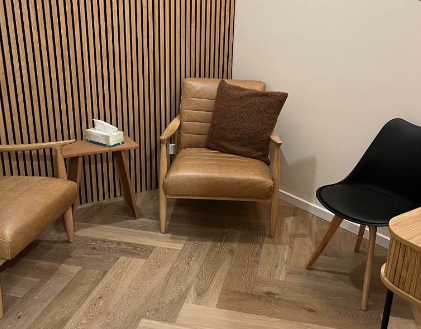 Waiting room with tan leather chairs, a brown pillow, a black chair, a side table with tissues, and wooden paneling on the wall.