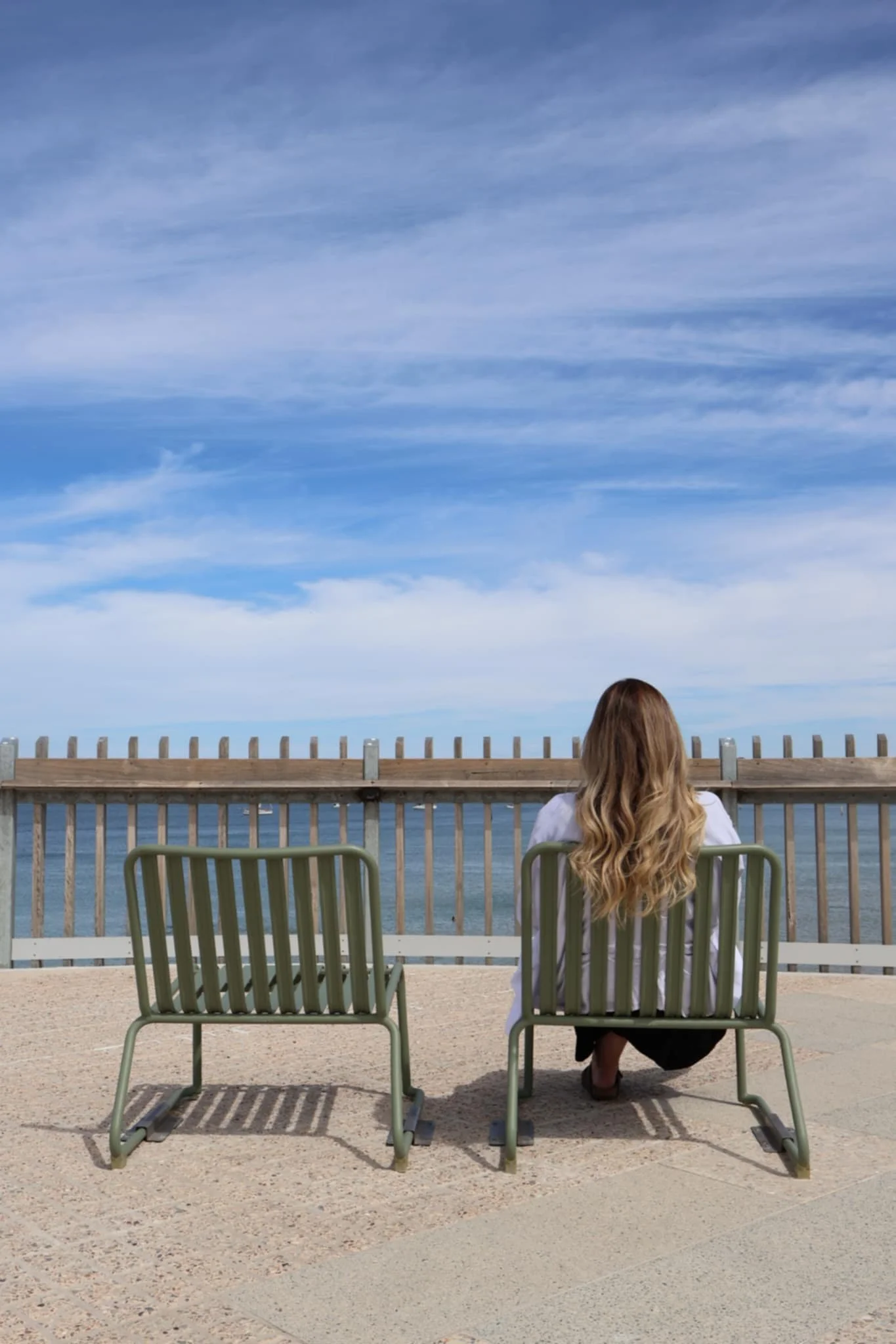 A woman with long, wavy hair is sitting on a metal lawn chair facing the ocean. In front of her, there are two empty similar chairs. She appears to be at a beach or seaside location, with a sandy ground, a wooden fence, and the ocean visible in the background under a partly cloudy sky.