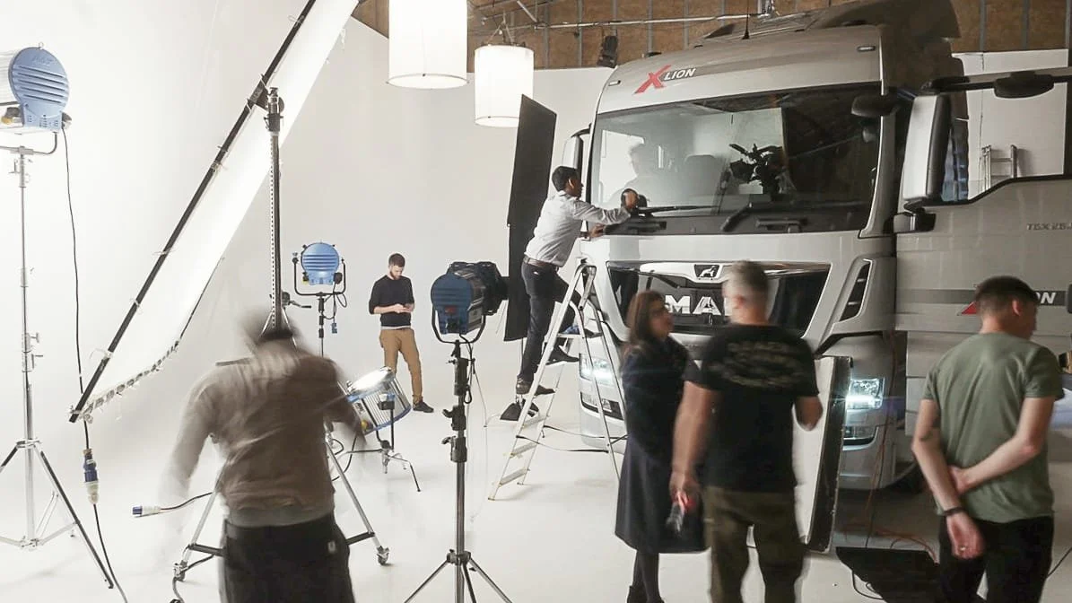 Film crew setting up equipment around a large gray truck with a boxy cab in a white studio space.