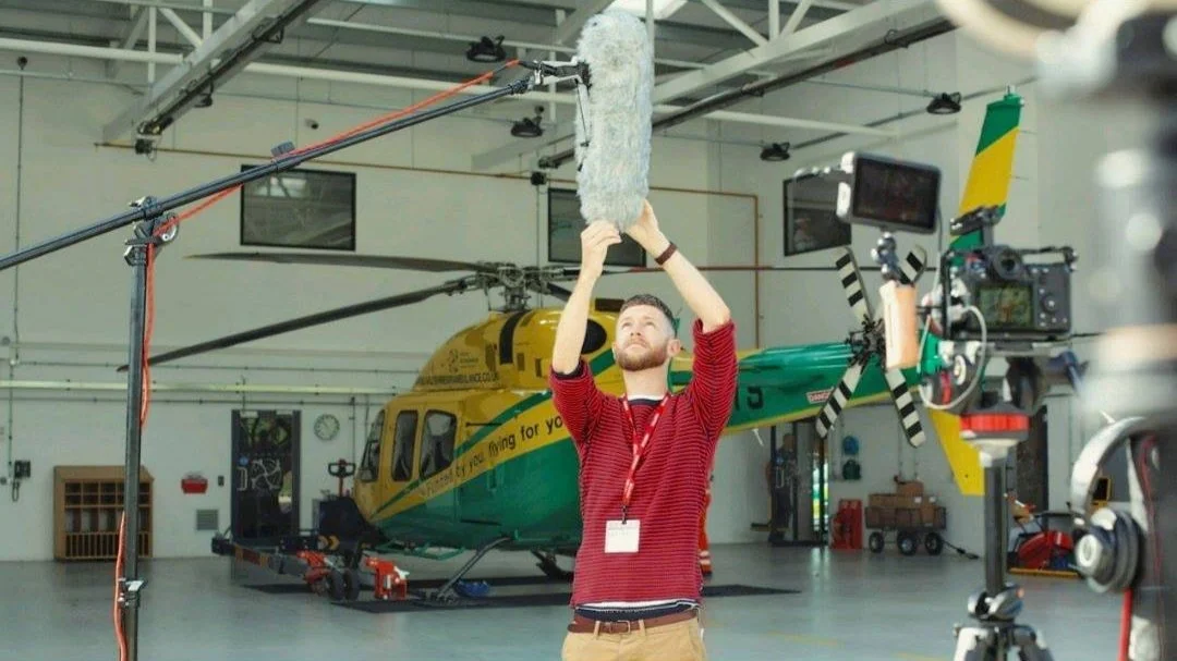A man wearing a red striped shirt adjusting a microphone boom pole inside a hangar with a helicopter in the background.