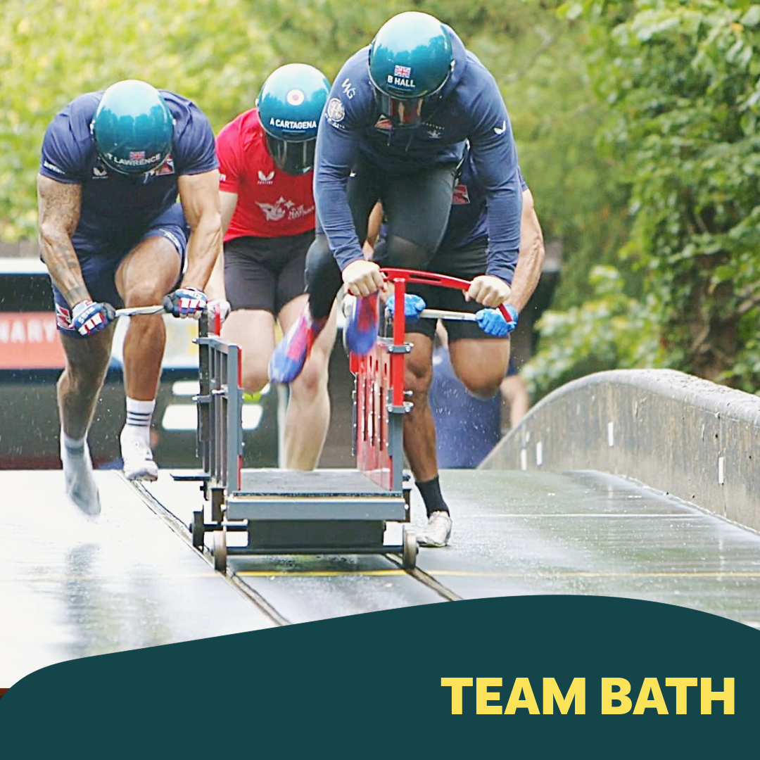 Three men in helmets and athletic gear pushing a bath across a track with a shower of water around them. The bath has a red handle and is part of a team activity.