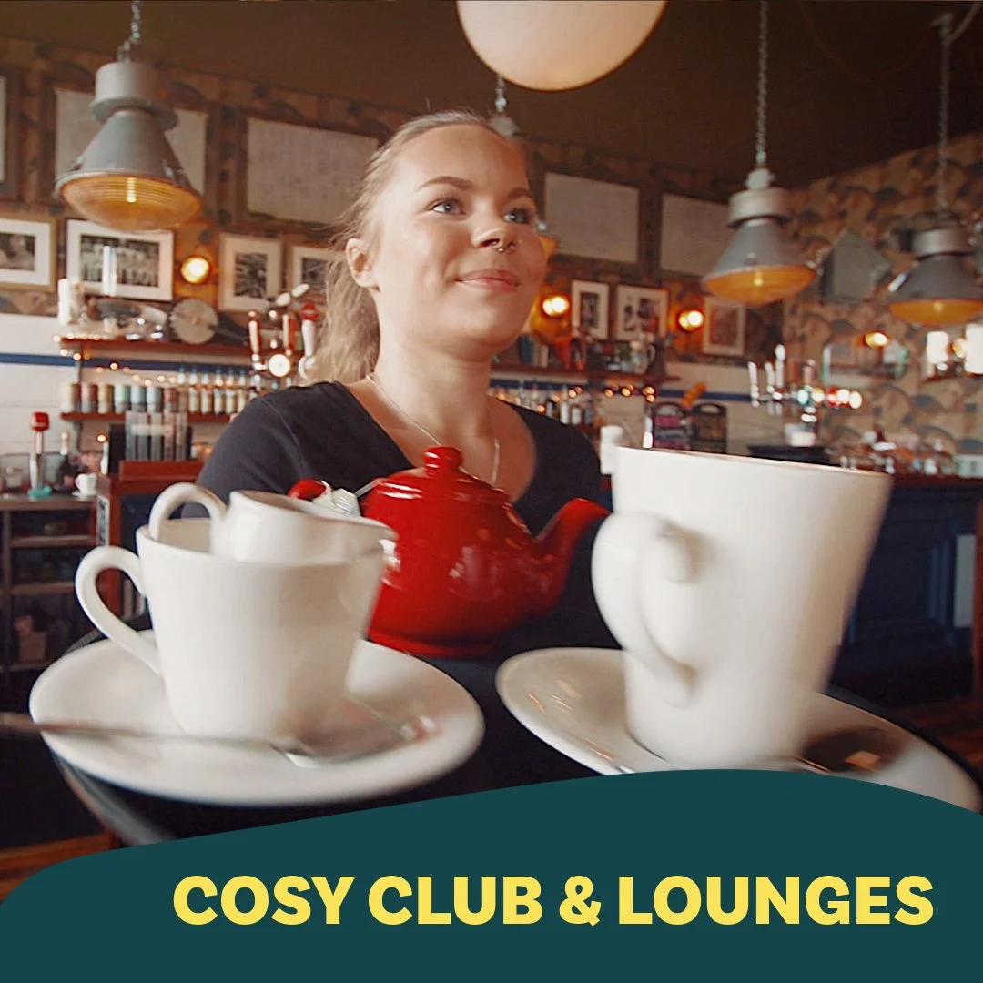 A young woman with a smiling expression sitting at a table in a cozy cafe, surrounded by a warm, inviting interior with framed photos and lights, with a red teapot and white cups on the table, and a promotional banner saying 'Cosy Club & Lounges' at the bottom.