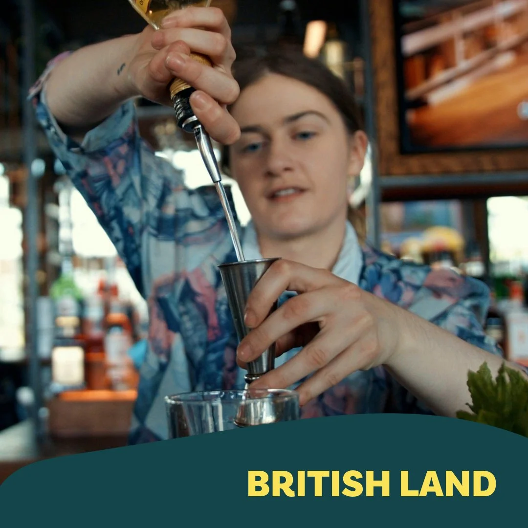 Person pouring liquid from a metal tool into a glass, with a background of a bar or restaurant setting, and the text 'British Land'.