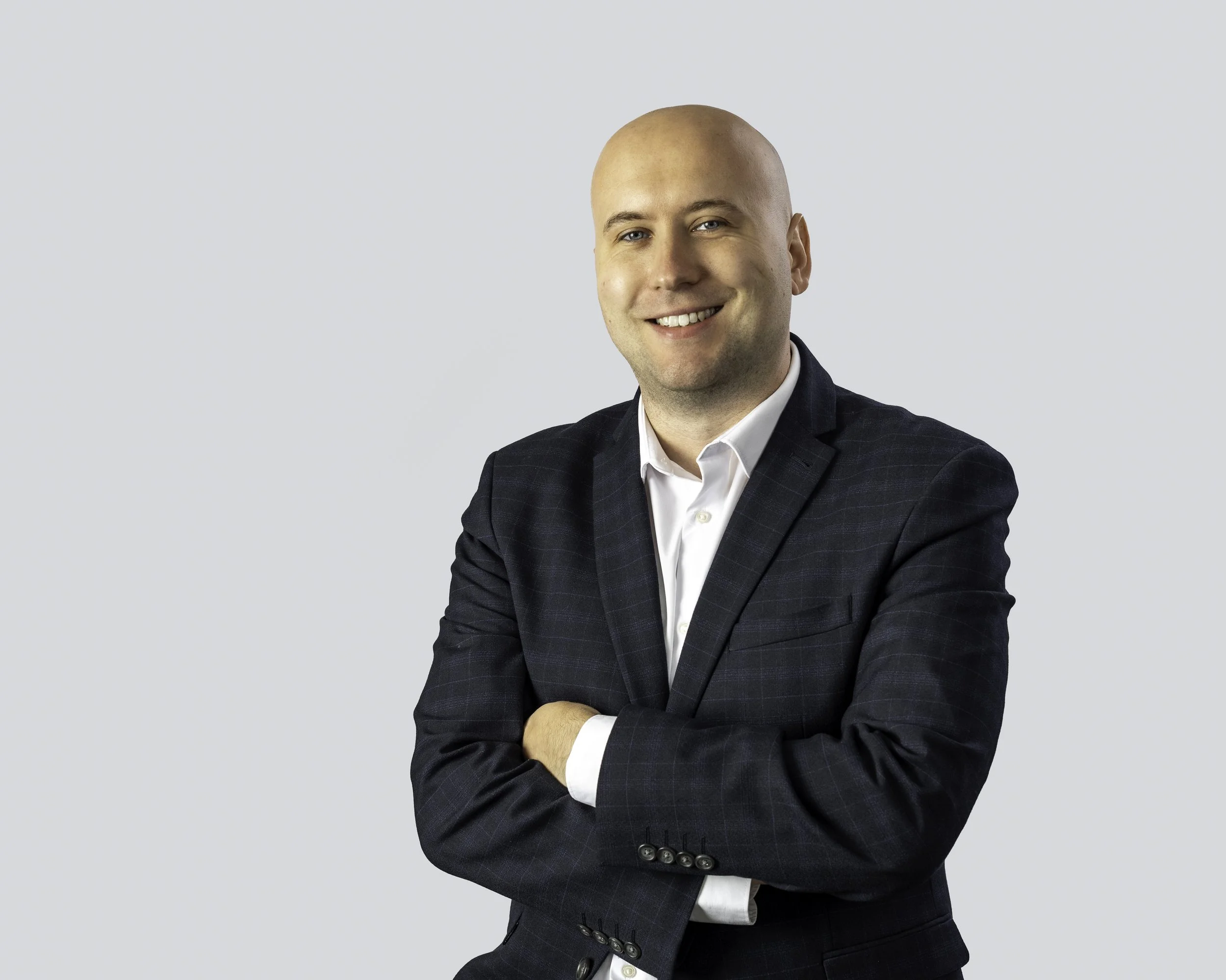 A bald man wearing a dark plaid suit with a white shirt, smiling with arms crossed, standing against a plain light background.