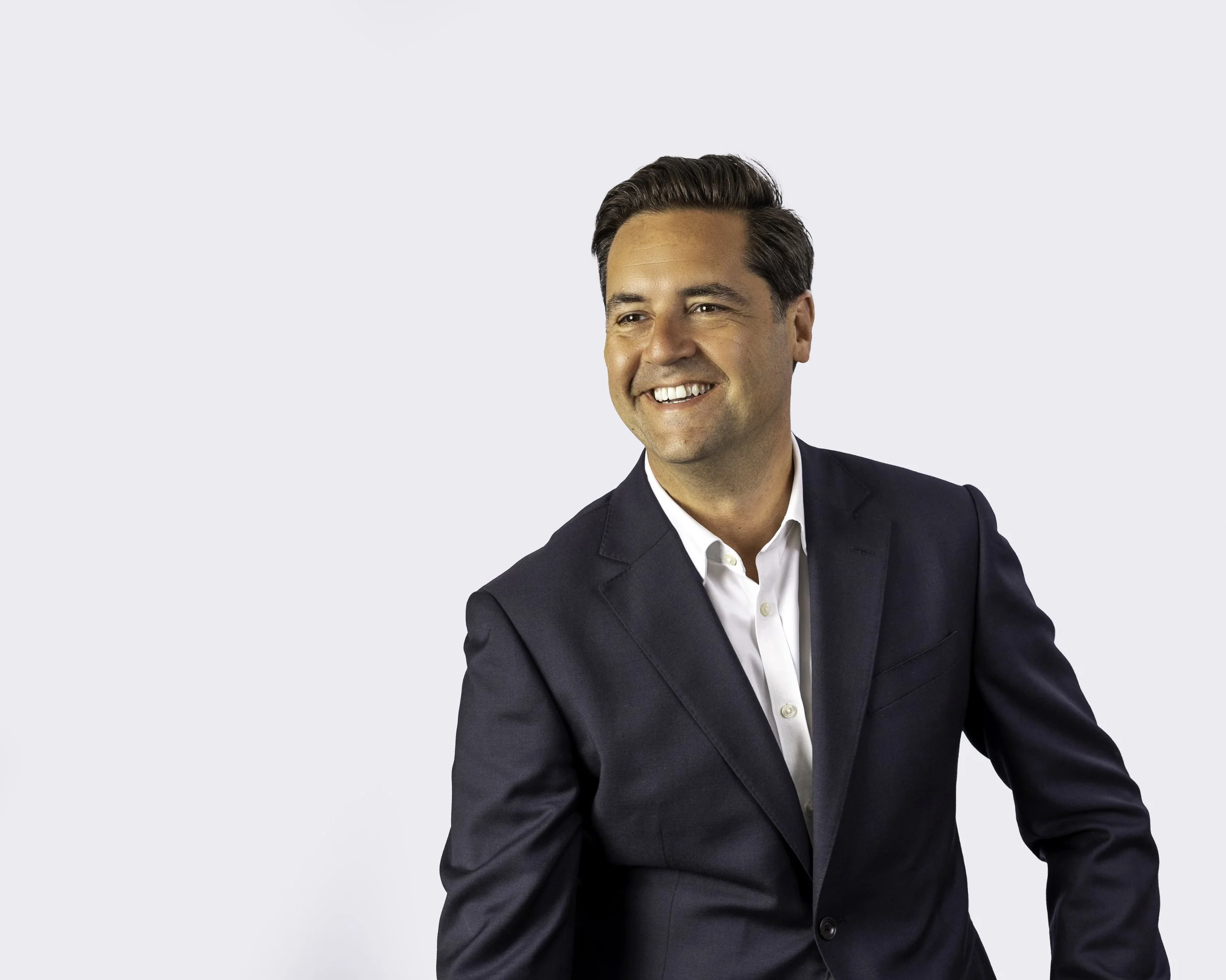 A man in a dark suit and white shirt smiling and looking to his left, against a plain light background.