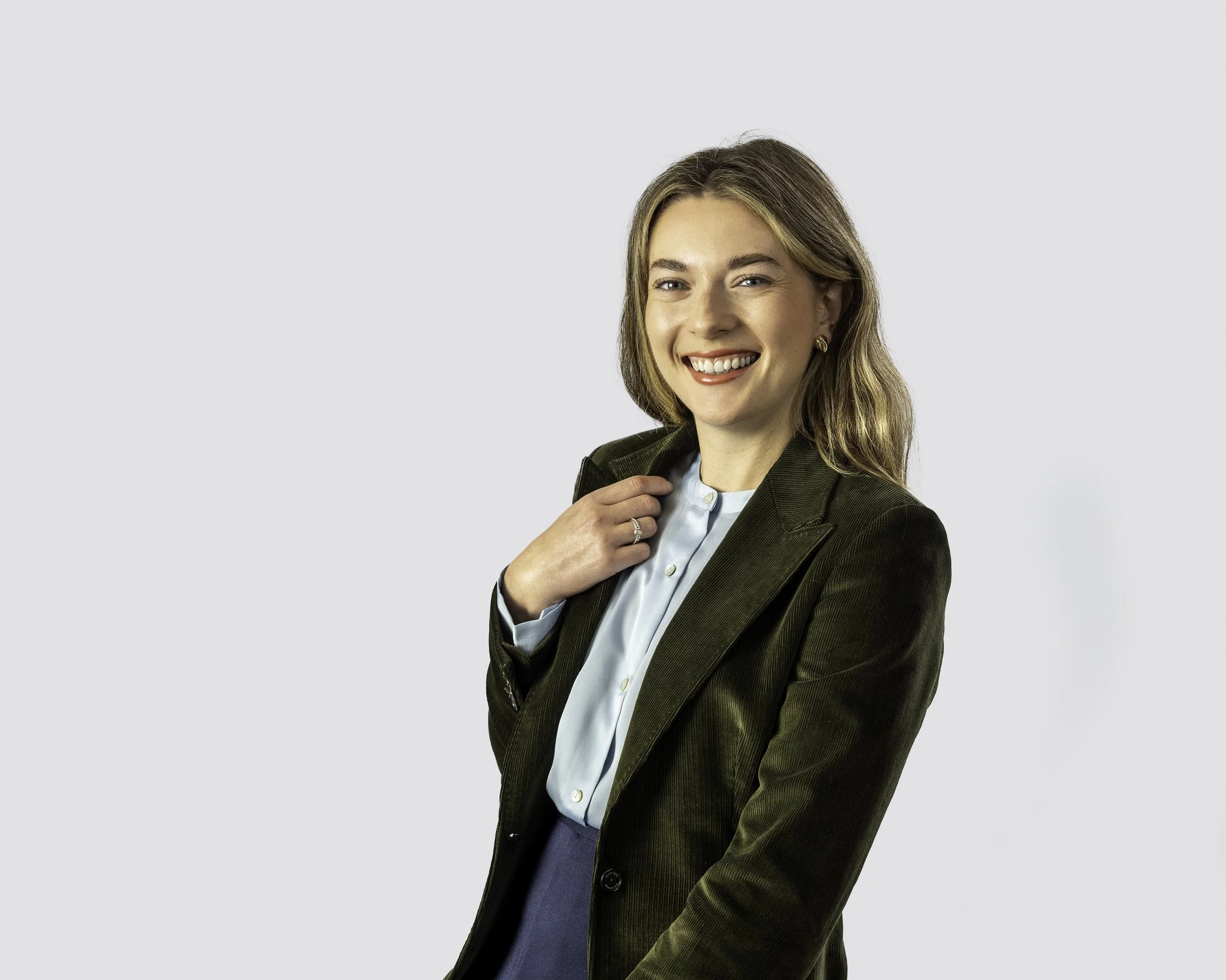 A smiling woman with wavy light brown hair, wearing a dark green blazer over a light blue blouse, against a plain light gray background.