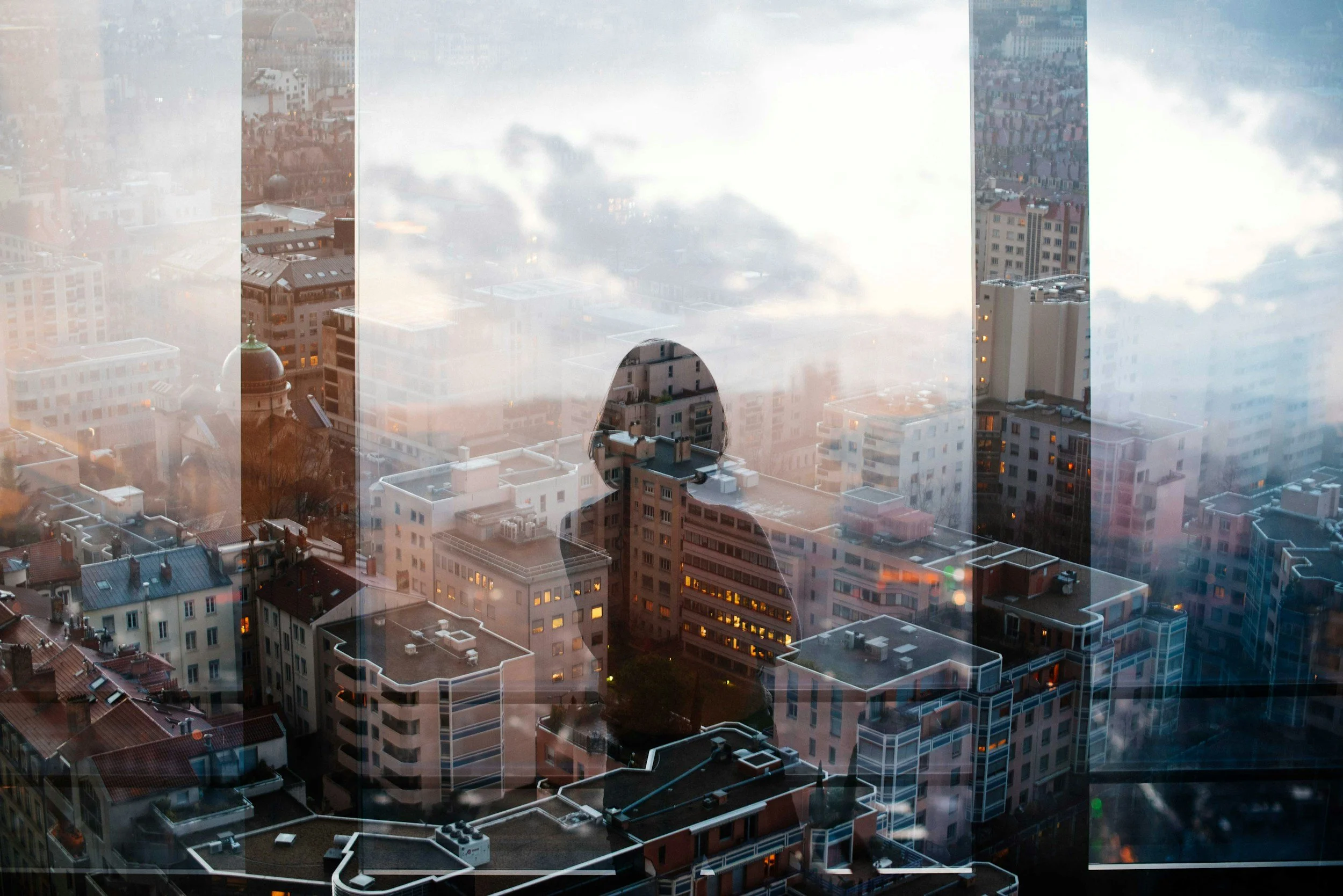 A cityscape view through a large glass window showing numerous high-rise buildings, some with antennas and rooftop equipment, under cloudy sky at dusk.