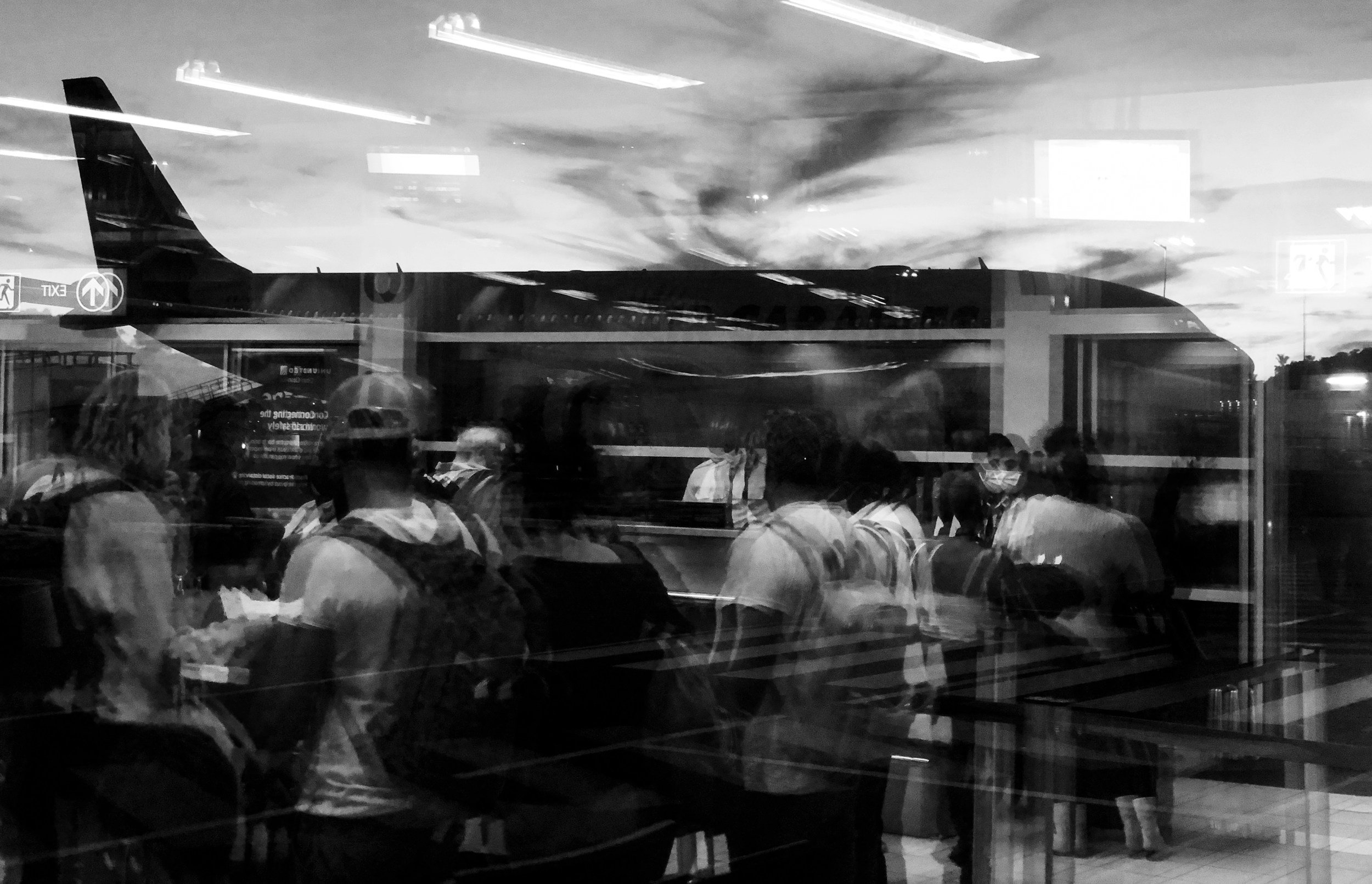 A view of an airplane through a glass window with reflections of people and lights inside a terminal, black and white photo.