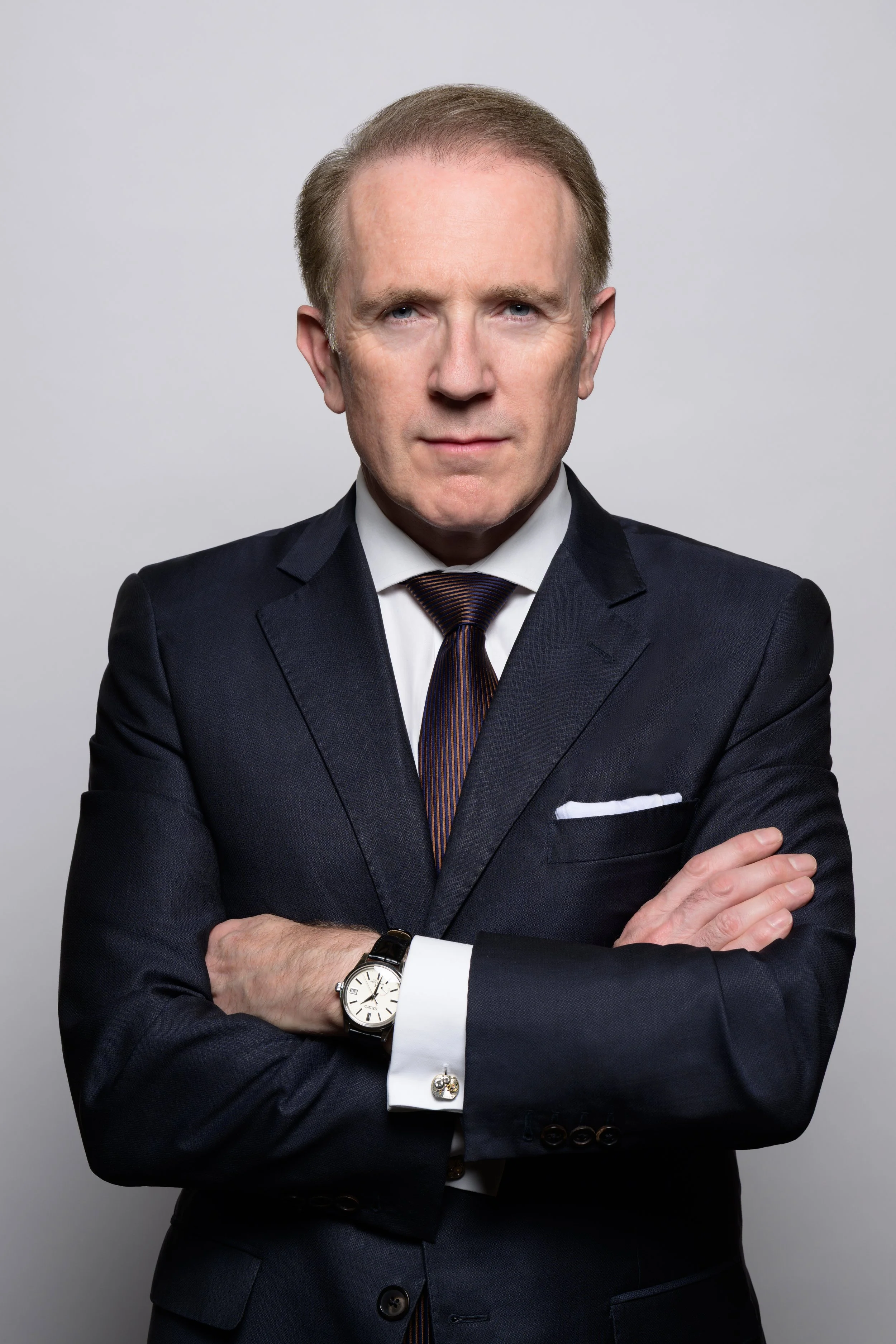 A man in a dark suit with a white shirt and a striped tie, wearing a wristwatch, standing with arms crossed against a plain light gray background.