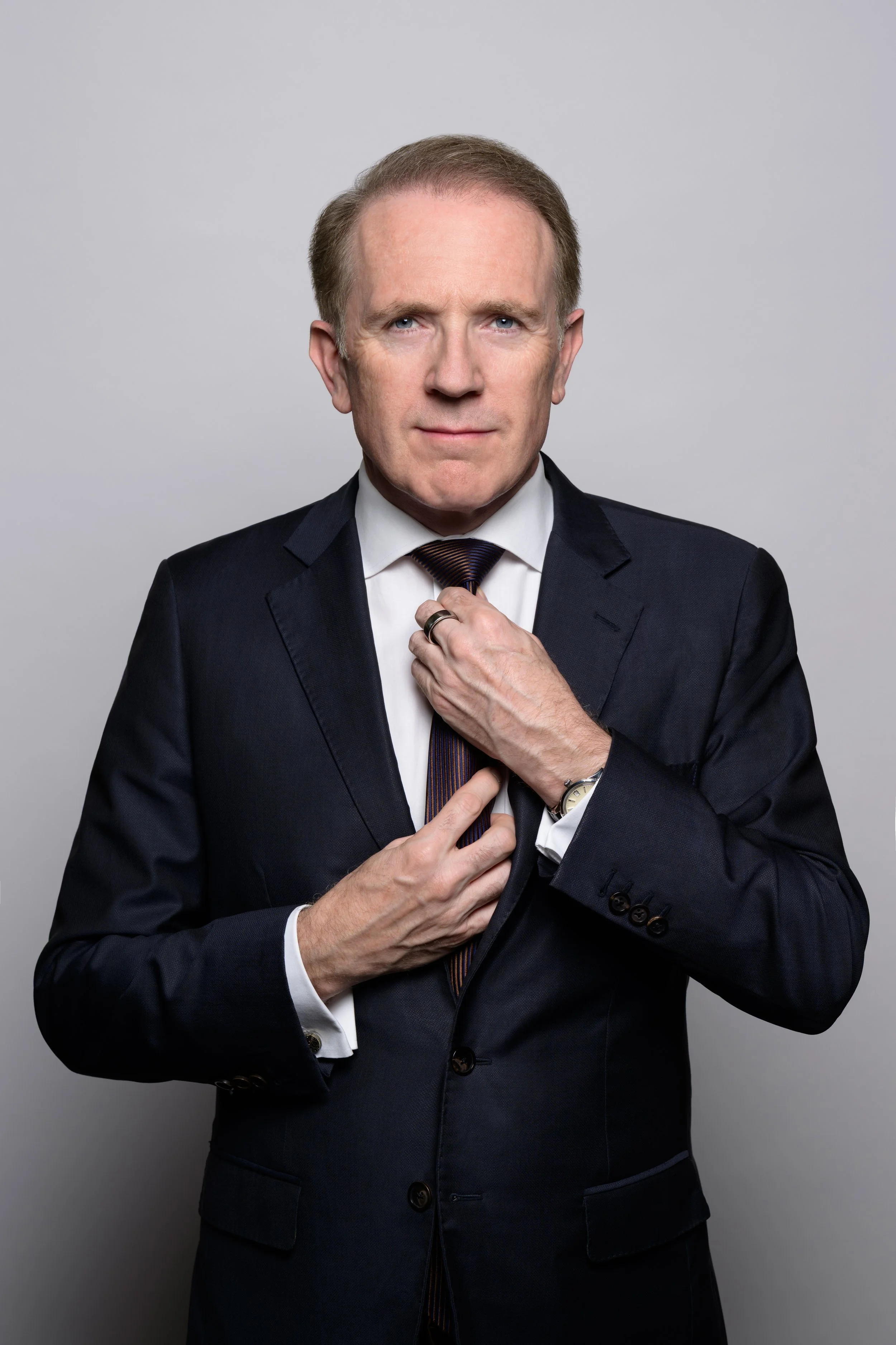 A middle-aged man with light skin, blue eyes, and short light brown hair, wearing a dark suit, white shirt, and striped tie. He is adjusting his tie with his left hand and has a silver watch on his left wrist, standing against a gray background.