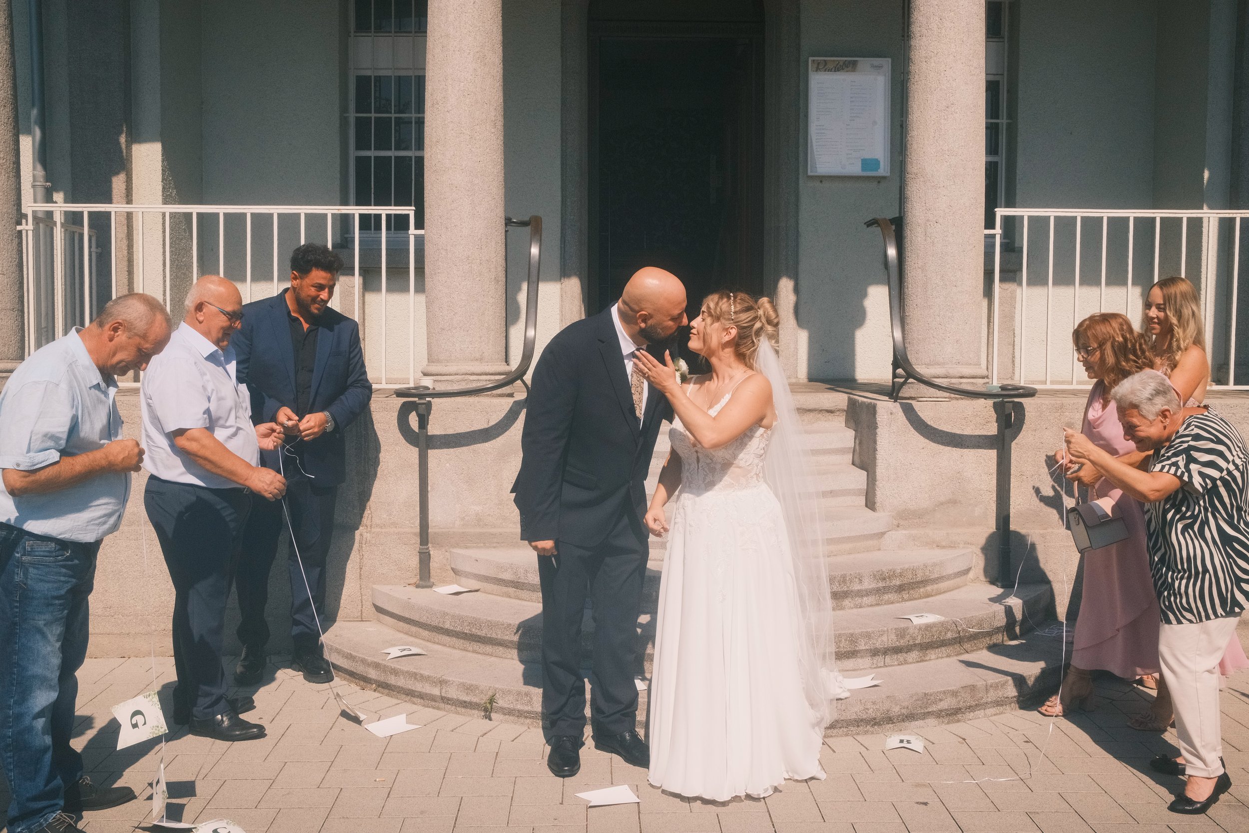 Brautpaar küsst sich vor dem Standesamt, Gäste knoten Papierschnipsel als Glücksbringer-Ritual, Hochzeit in Leipzig