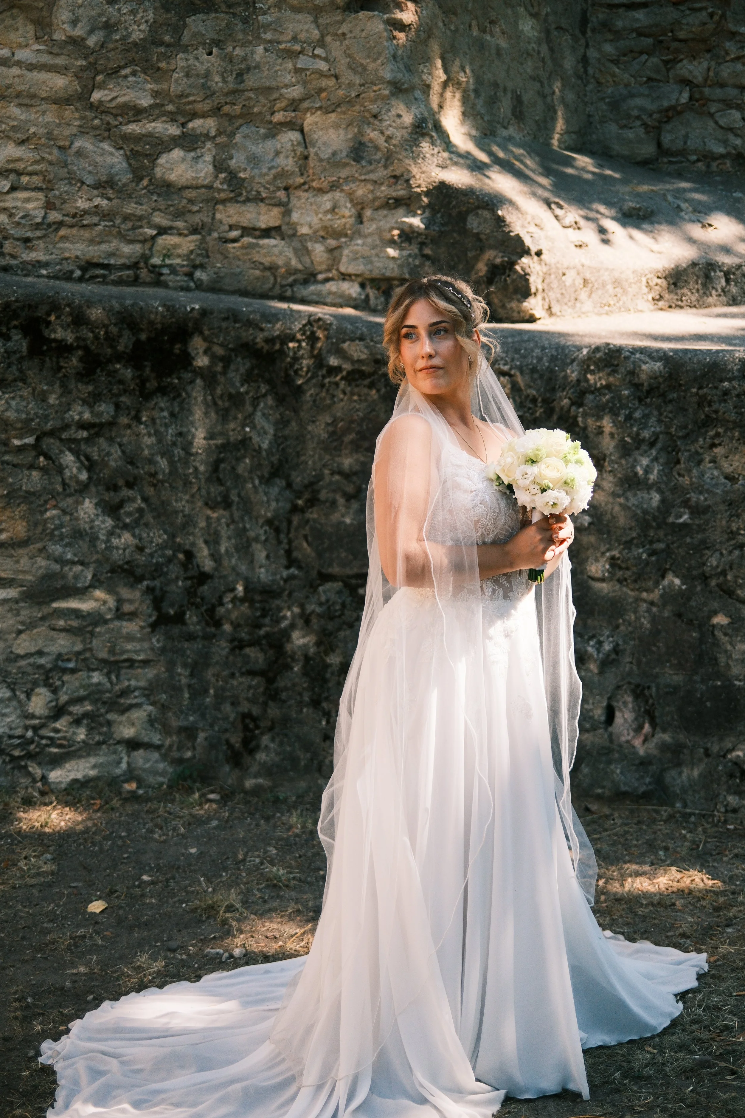 Lächelnde Braut im Vordergrund, Bräutigam wartet im Hintergrund vor einer Natursteinmauer, Hochzeit in Leipzig
