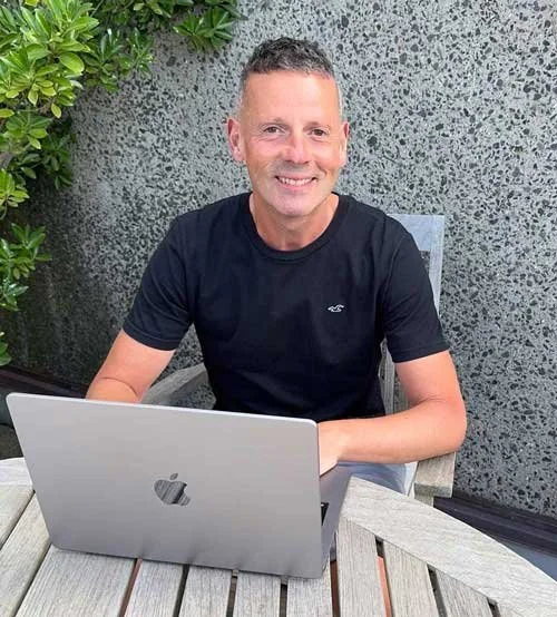 The author, Graeme Gordon, sitting outside at a table with a silver laptop, smiling, with green plants and a concrete wall in the background.