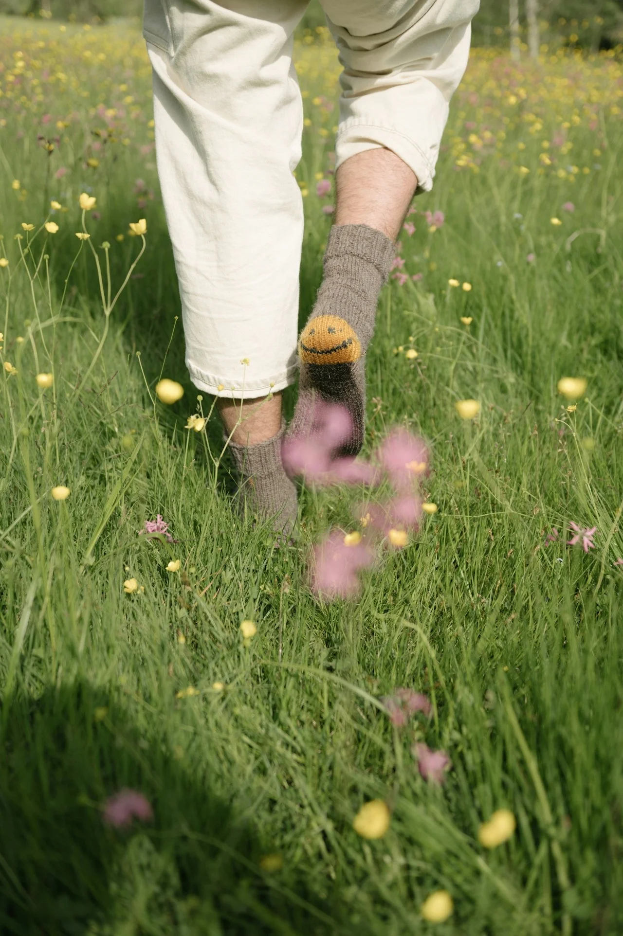 Close-up of a person walking through a field of yellow, pink, and purple wildflowers, wearing beige pants and a gray sock with a yellow smiley face.
