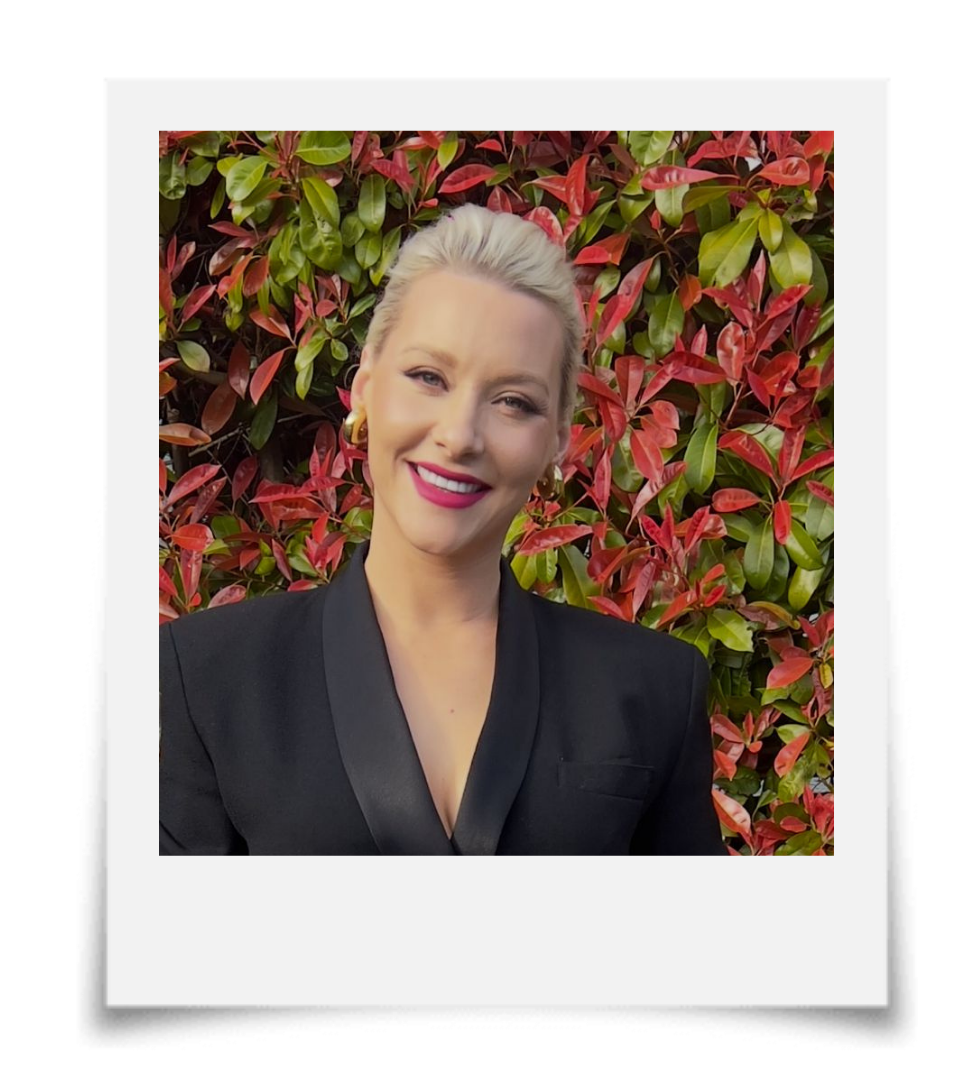 A woman with blonde hair styled in an updo, wearing a black blazer and gold hoop earrings, smiling in front of a red and green leafy background.