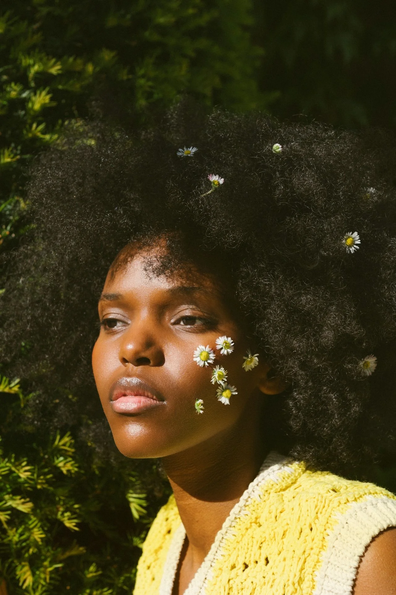 Close-up of a woman with dark skin and voluminous curly hair with small daisies woven into her hair and face, wearing a yellow knitted top, outdoors with greenery in the background.