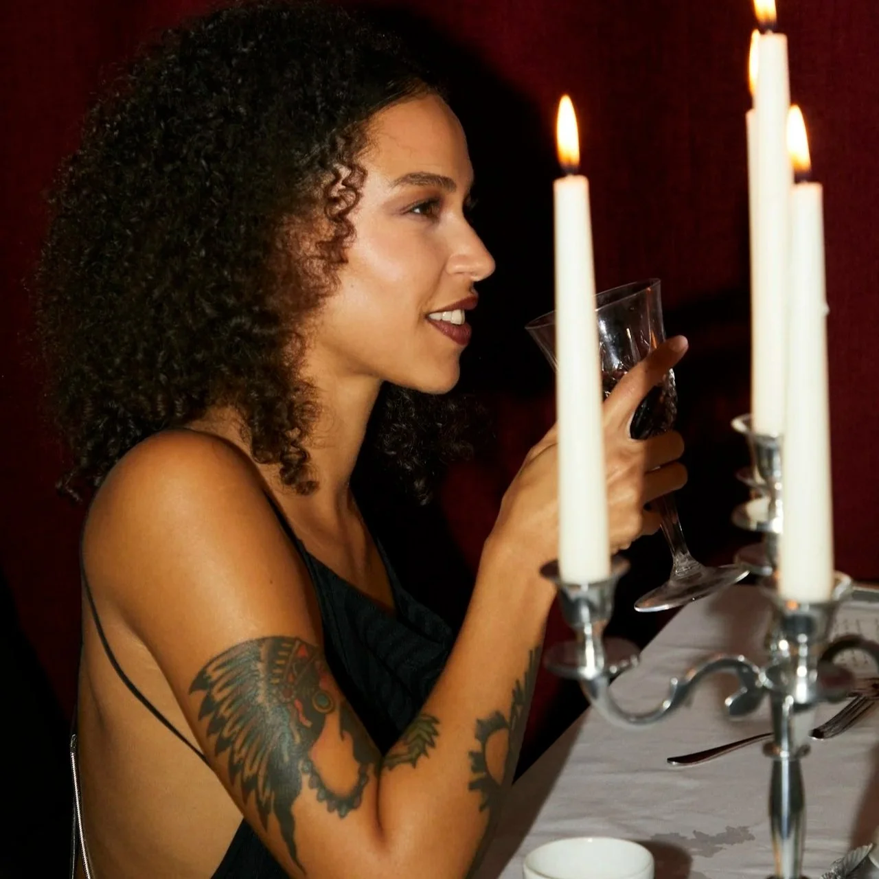 A woman with curly hair and tattoos on her arm holding a glass of red wine at a dinner table with white candles in a candelabrum.