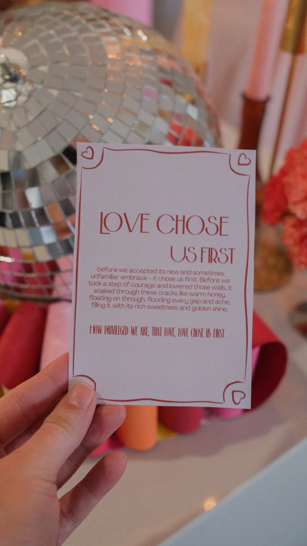 A person holding a white card with red text and decorative border that reads, "Love chose us first". In the background, there's a reflective, mosaic-style lamp and blurred colorful surroundings.