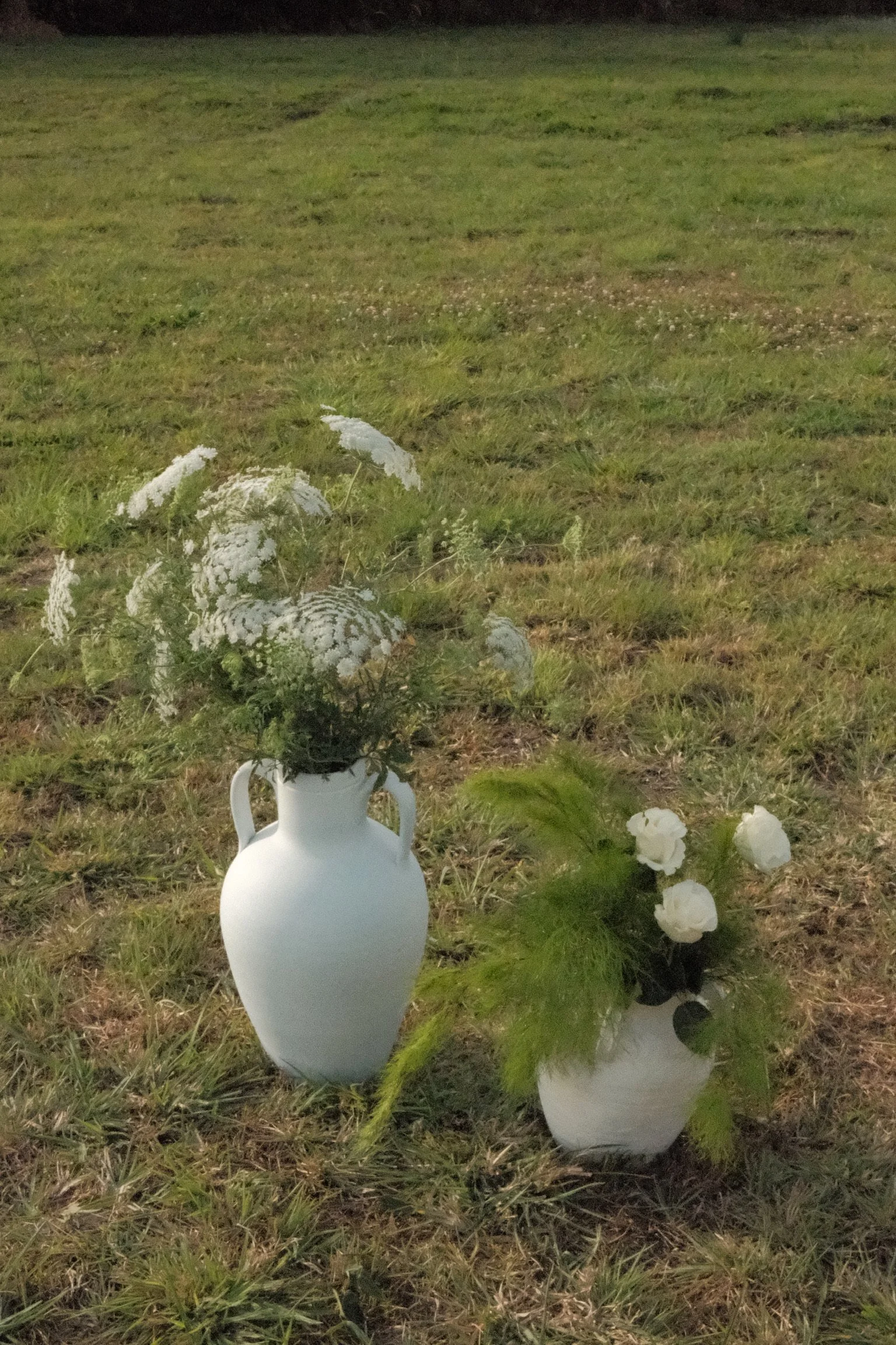 A white ceramic vase filled with white and green flowers is placed on the grass in an outdoor setting, with grass and a dark background in the distance.