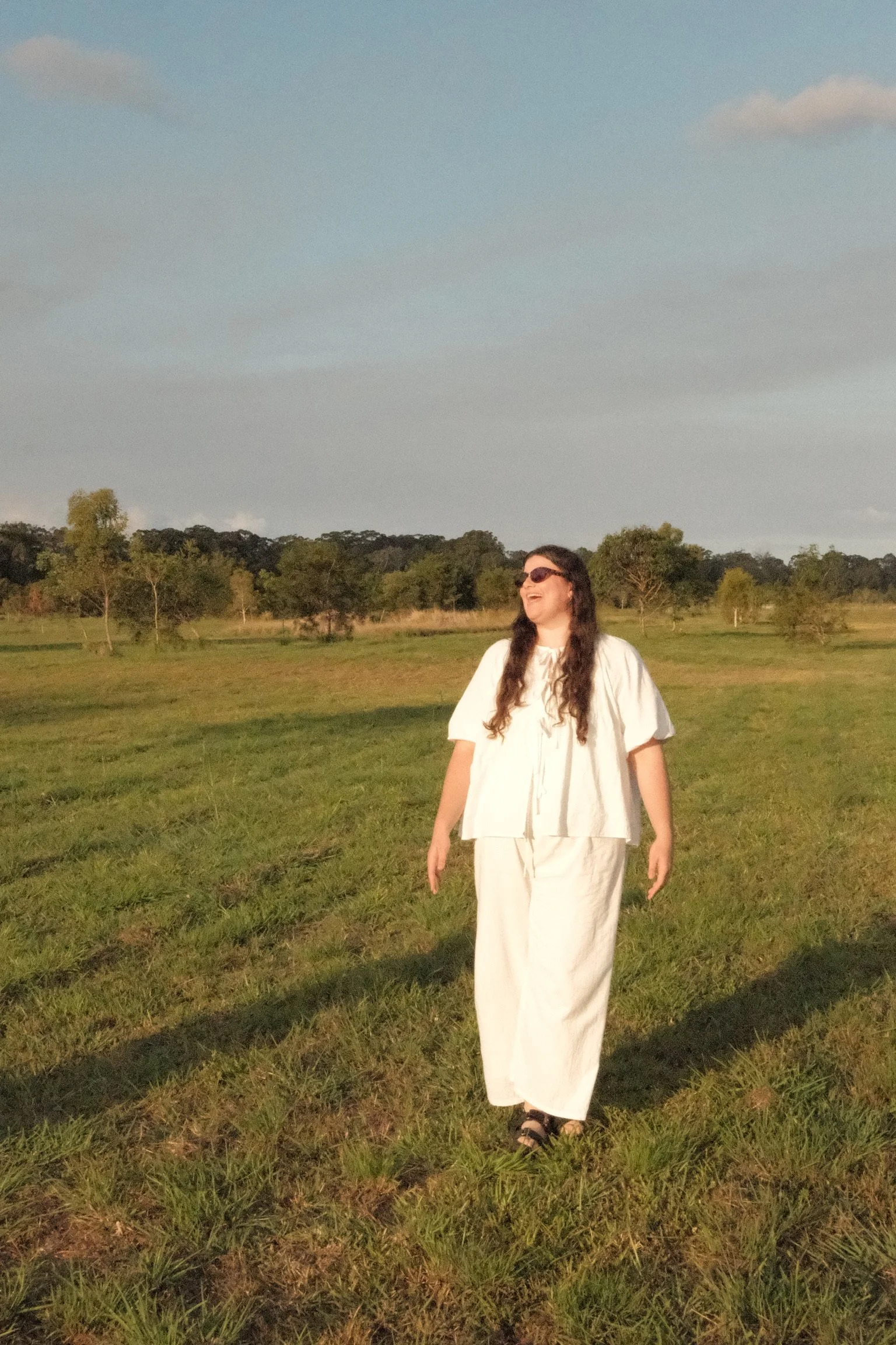 A woman with long dark hair, wearing sunglasses, a white top, and white pants, walking on grass in an open field during the daytime, smiling and looking to her left, with trees and a clear sky in the background.