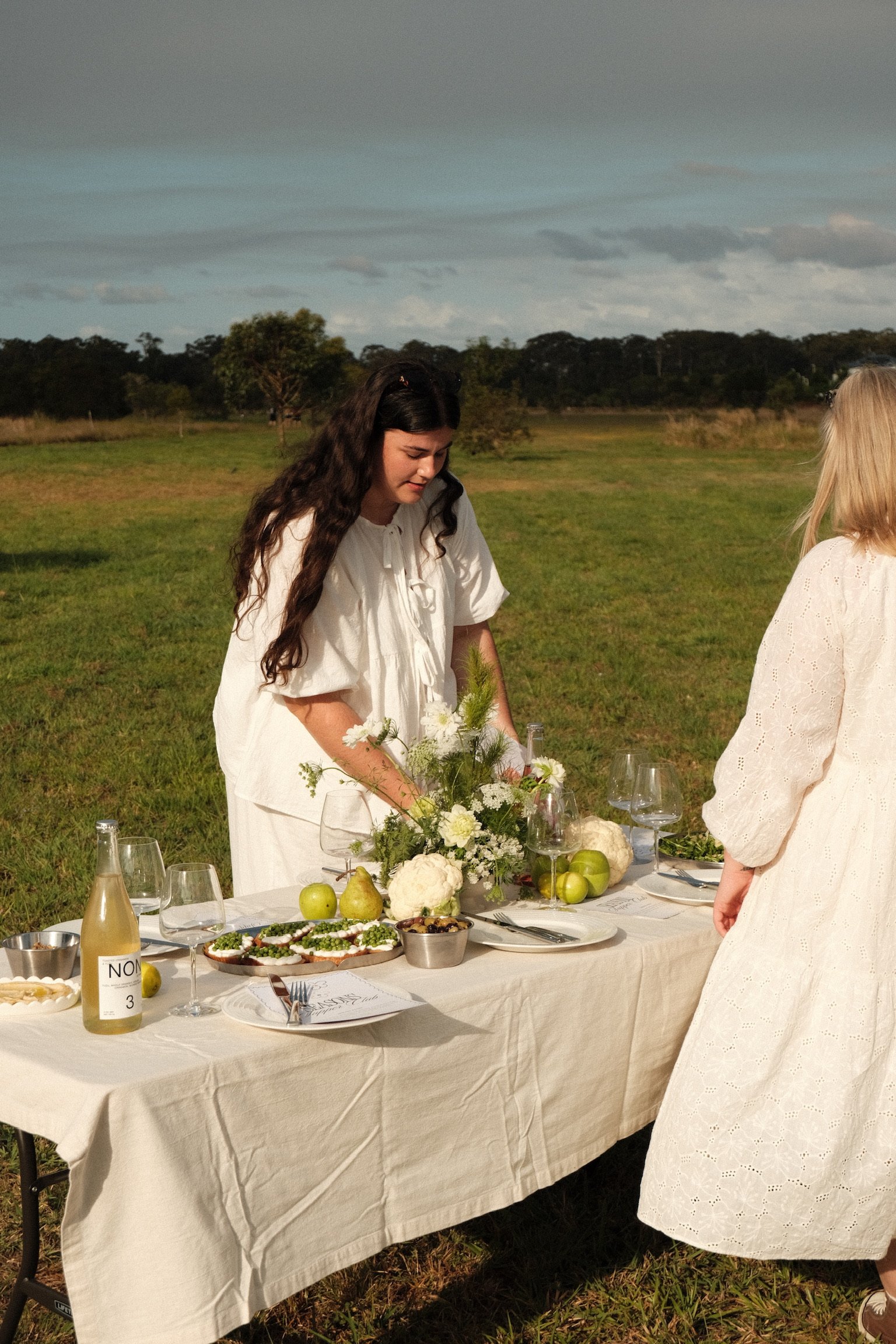 Two women in white dresses are setting a table outdoors in a grassy field with trees and a cloudy sky in the background. The table is decorated with flowers, apples, cauliflower, and various foods and drinks.