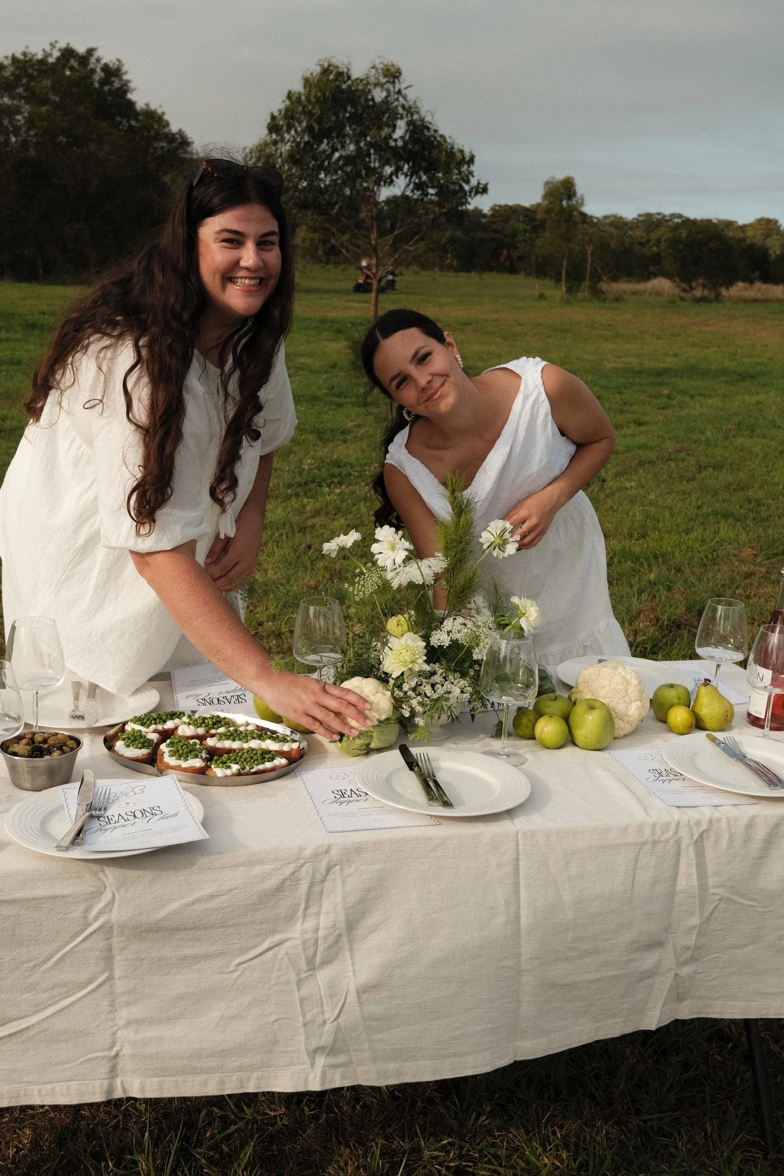Two women in white dresses smiling at a table with food and flowers outdoors during the daytime.
