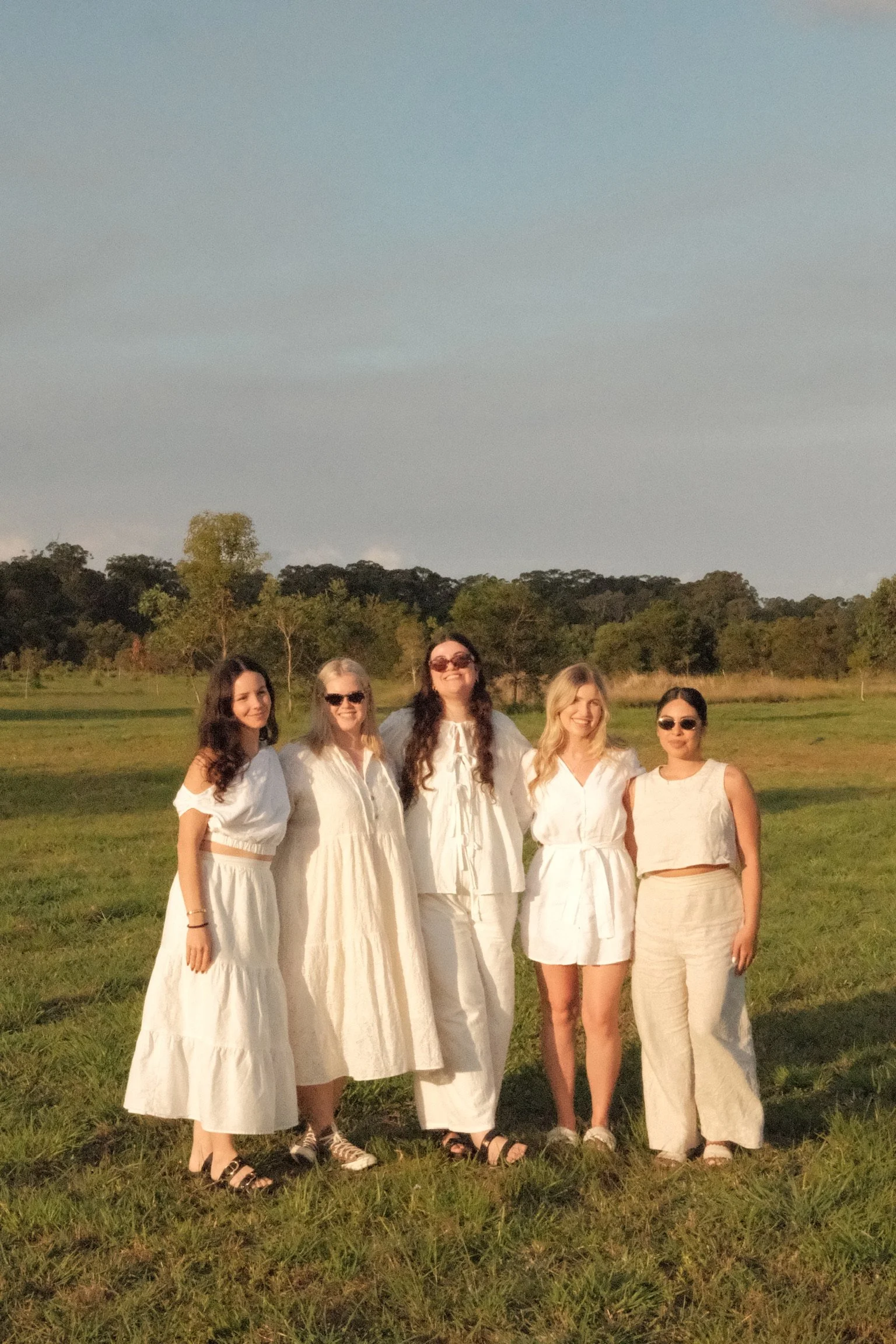 Six women standing outdoors on grass with trees and an open sky in the background, dressed in white clothing, some wearing sunglasses, smiling and posing for the camera during a sunny day.