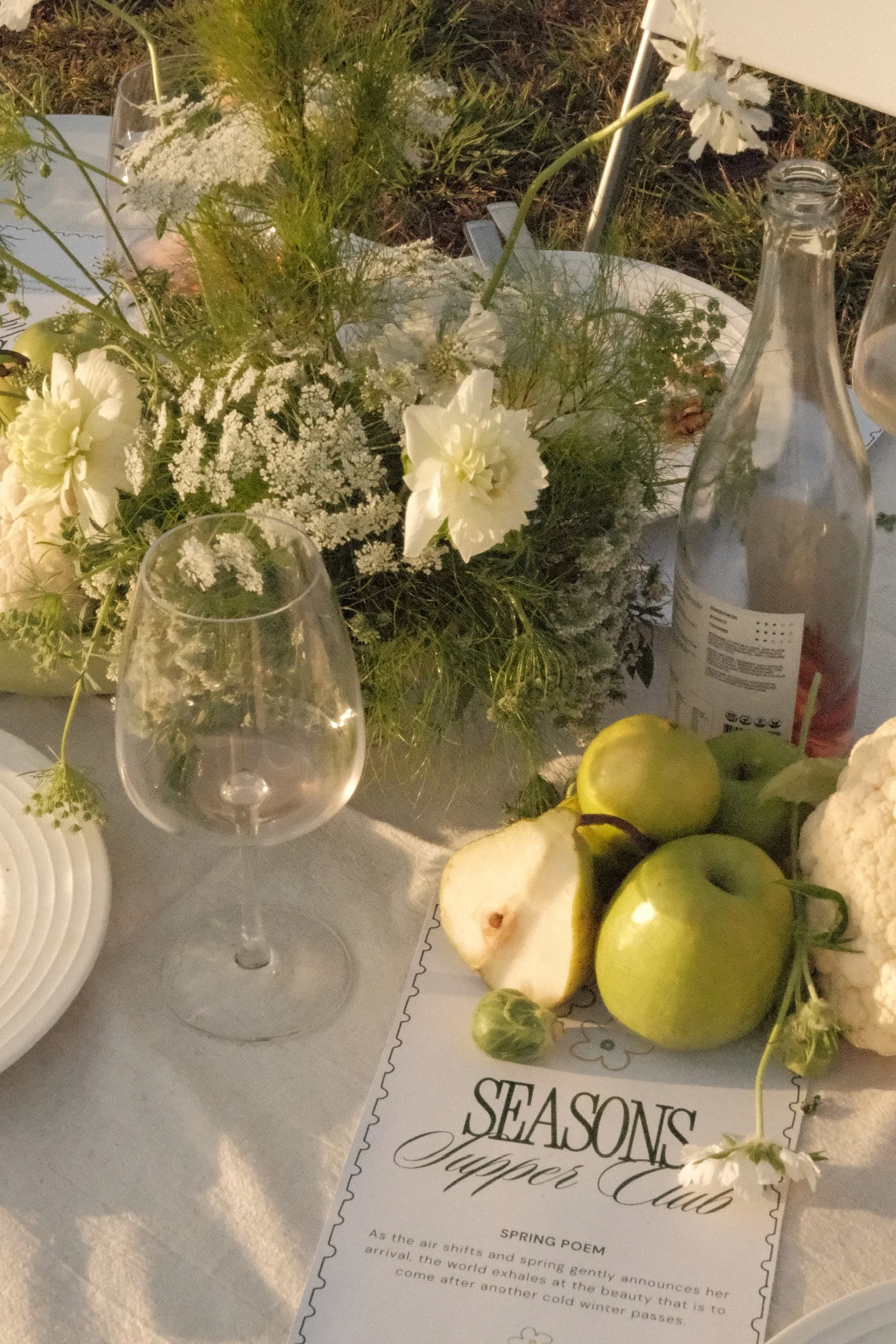 A table setting with a flower arrangement, a wine glass, and a bottle of sparkling water. There are apples and a sliced pear on the table, along with a menu titled 'Spring Poem' from the Seasons Tapper Club.