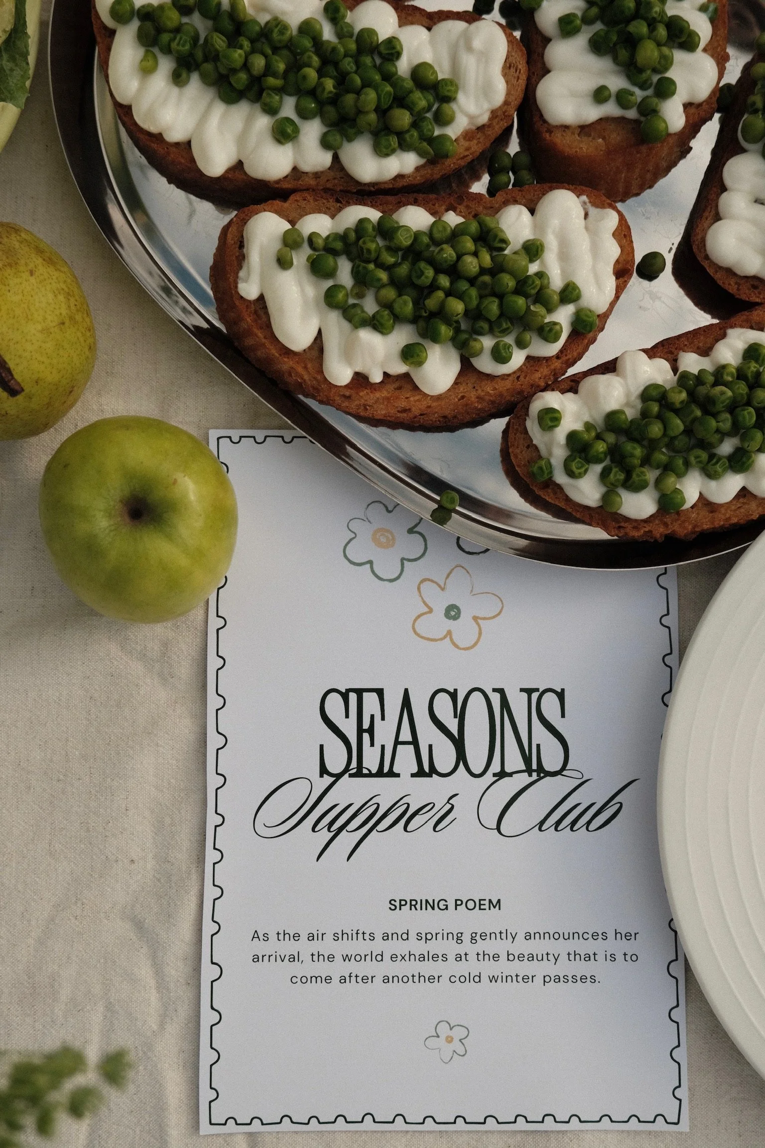 A silver tray with slices of bread topped with white cream cheese and green peas. Two green apples are placed beside a printed paper with the text 'SEASONS Summer' and a partial paragraph about spring.