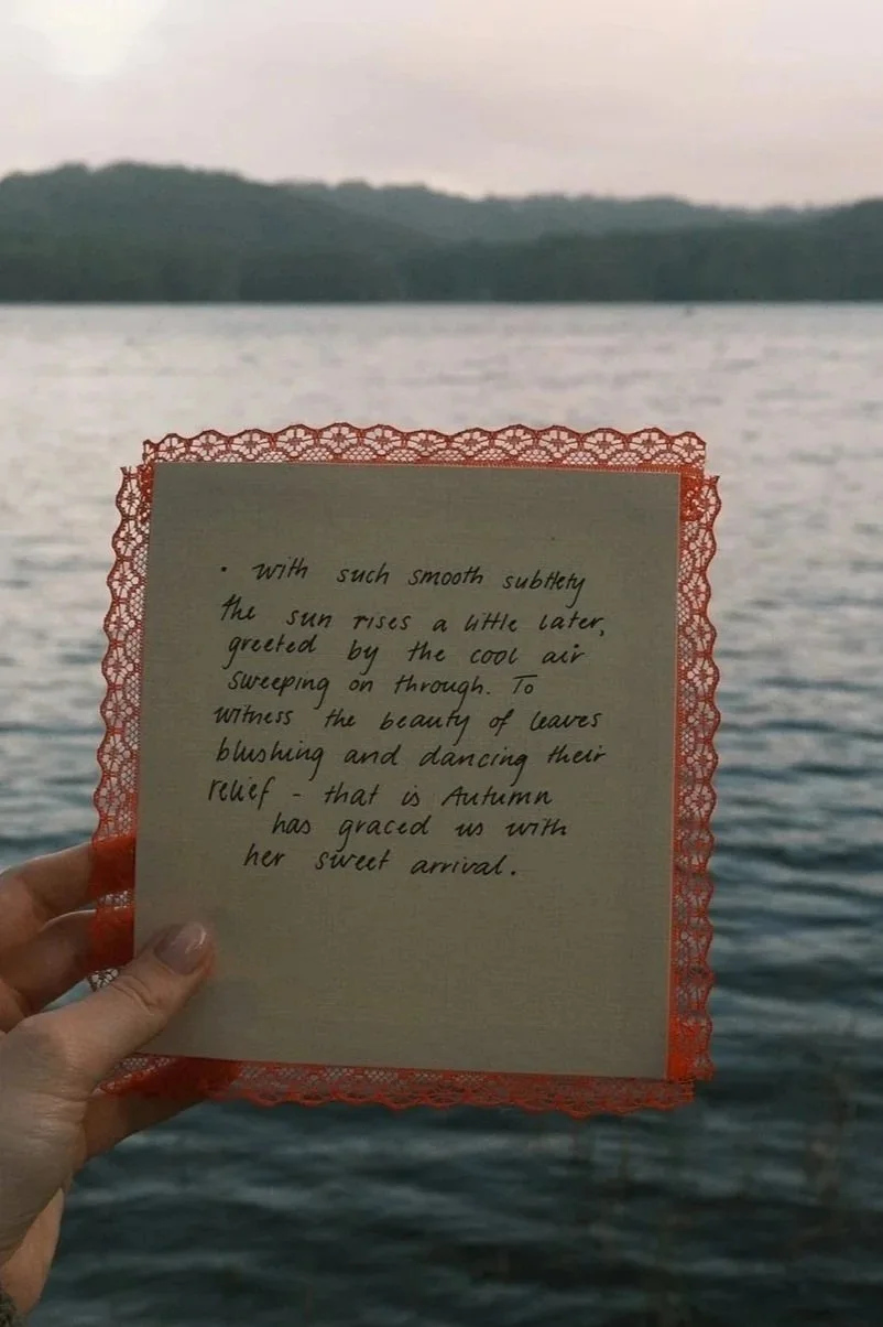 Hand holding a decorative notecard with handwritten text in front of a lake and distant forested shoreline during dusk.