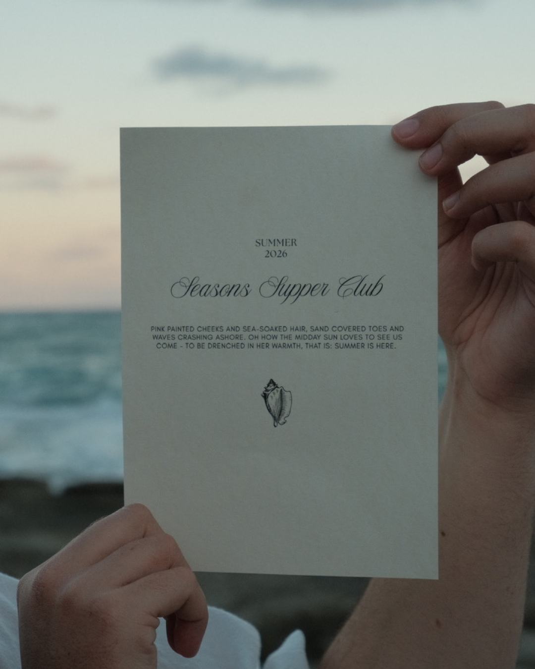 Person holding a printed invitation or program with the text 'Summer 2026 Seasons Supper Club' and a seashell illustration, at the beach during sunset or sunrise.