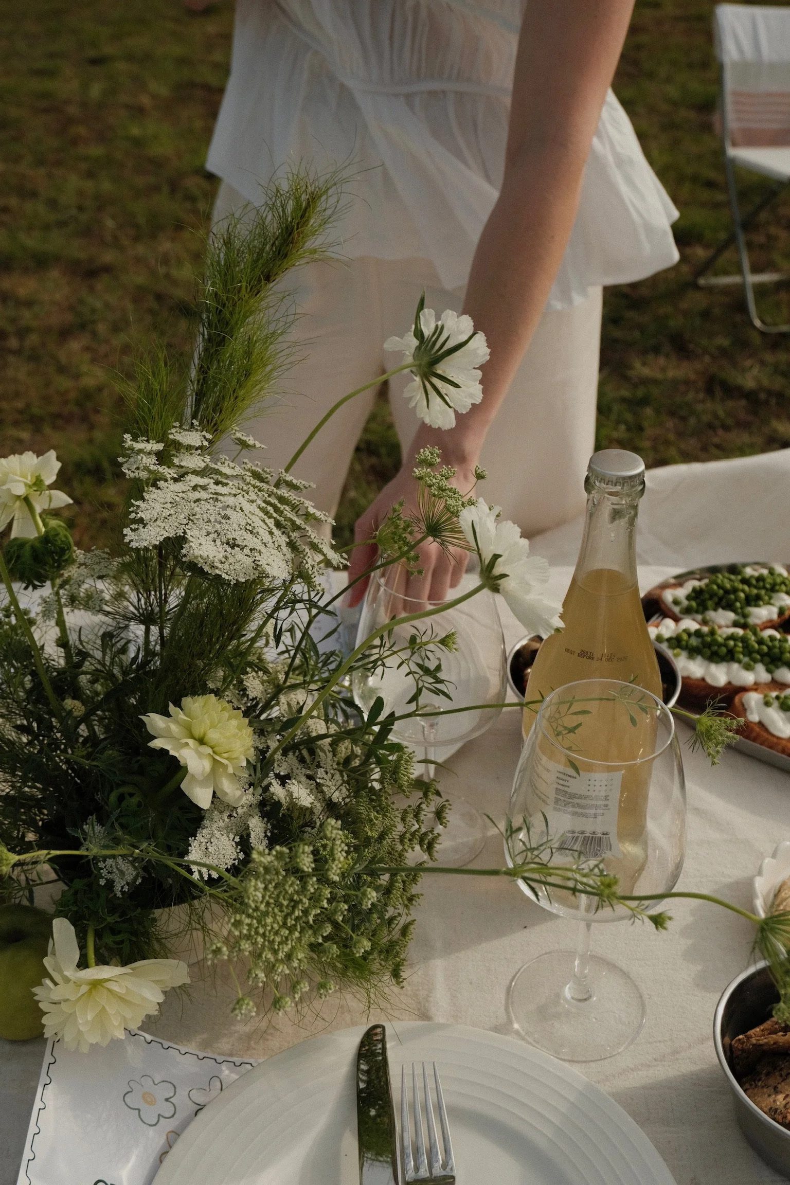 A white tablecloth with a floral pattern, decorated with a bouquet of white and green flowers and greenery. There are plates, glasses, and a bottle of sparkling beverage on the table. A person's hand is reaching across the table, with a white dress and outdoor setting visible.