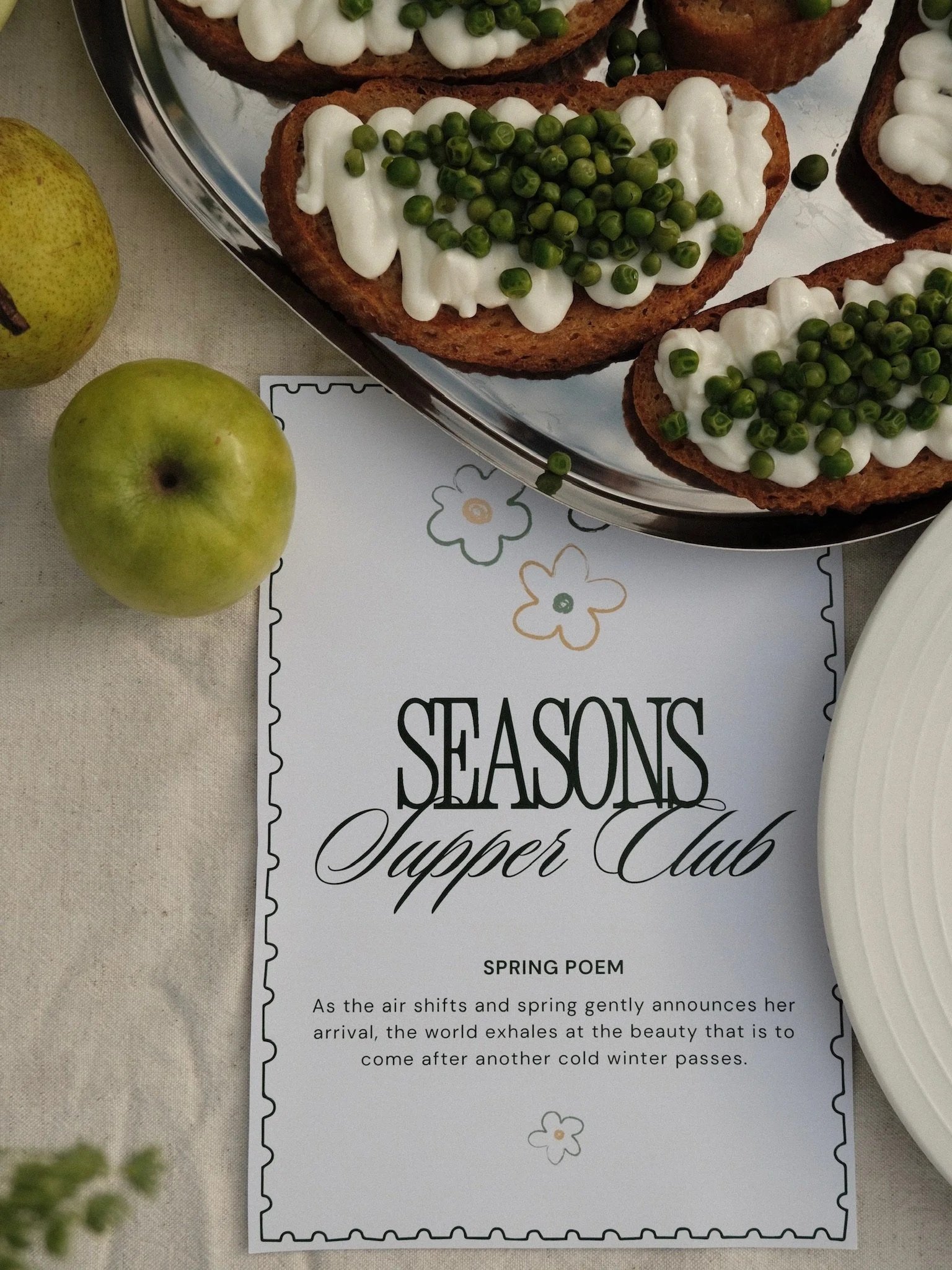 Close-up of a tray with slices of bread topped with white creamy spread and green peas, next to two green apples on a beige tablecloth, and a paper with the text 'SEASONS Suppper Club SPRING POEM'.