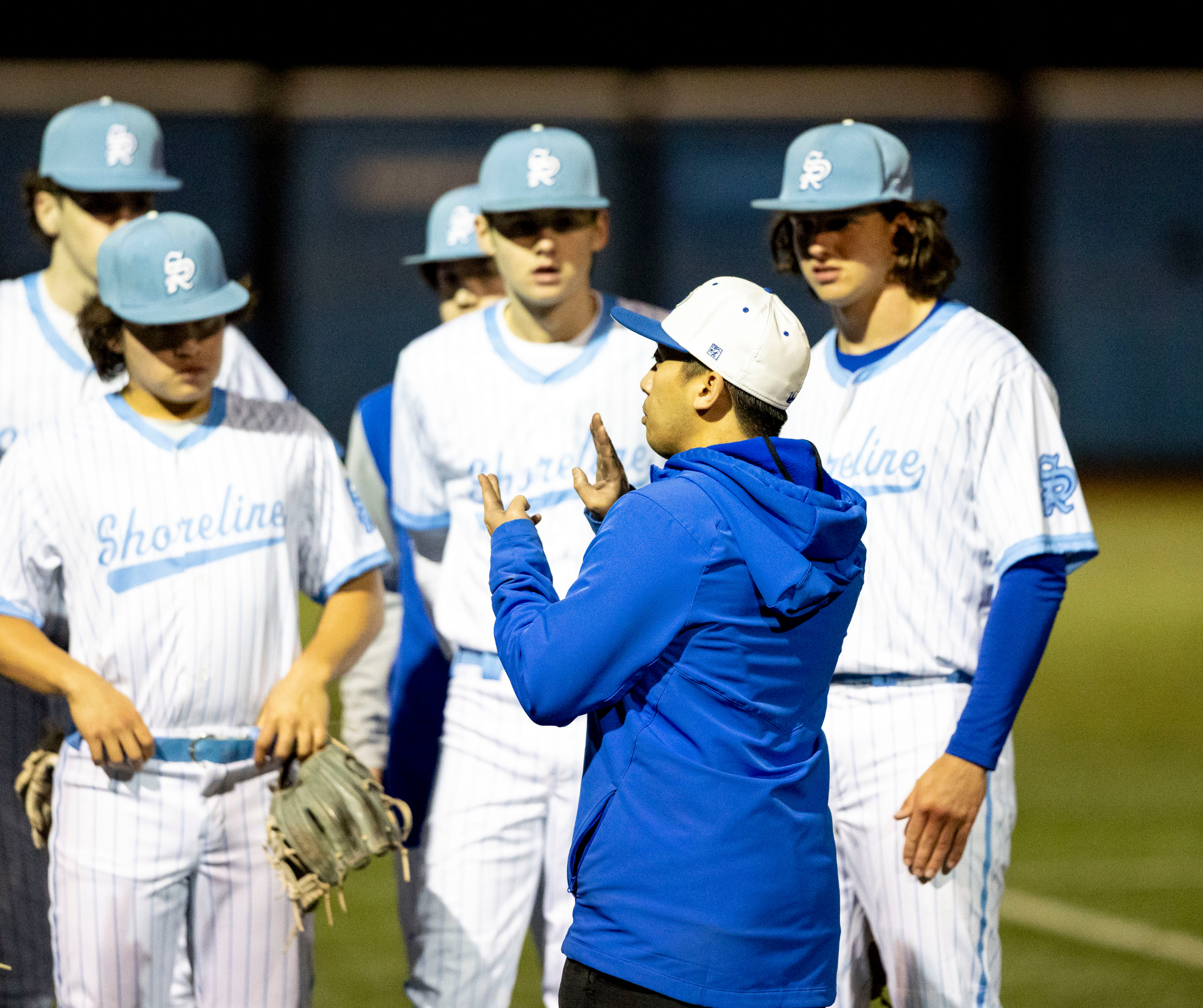 Baseball team members in blue and white uniforms and caps standing in a group, with one person in a blue jacket or hoodie talking to them during a game.