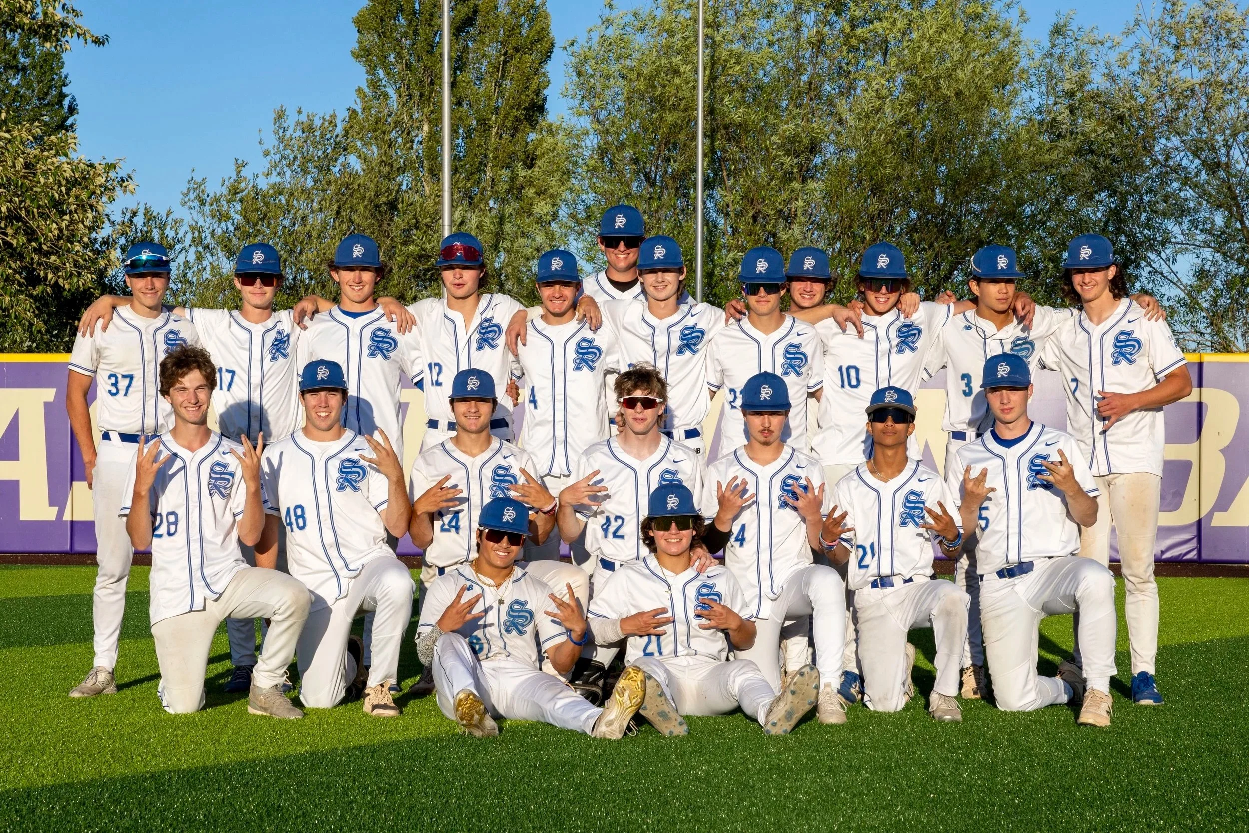 A group of young baseball players in uniform, posing together on a baseball field, smiling and making hand gestures.