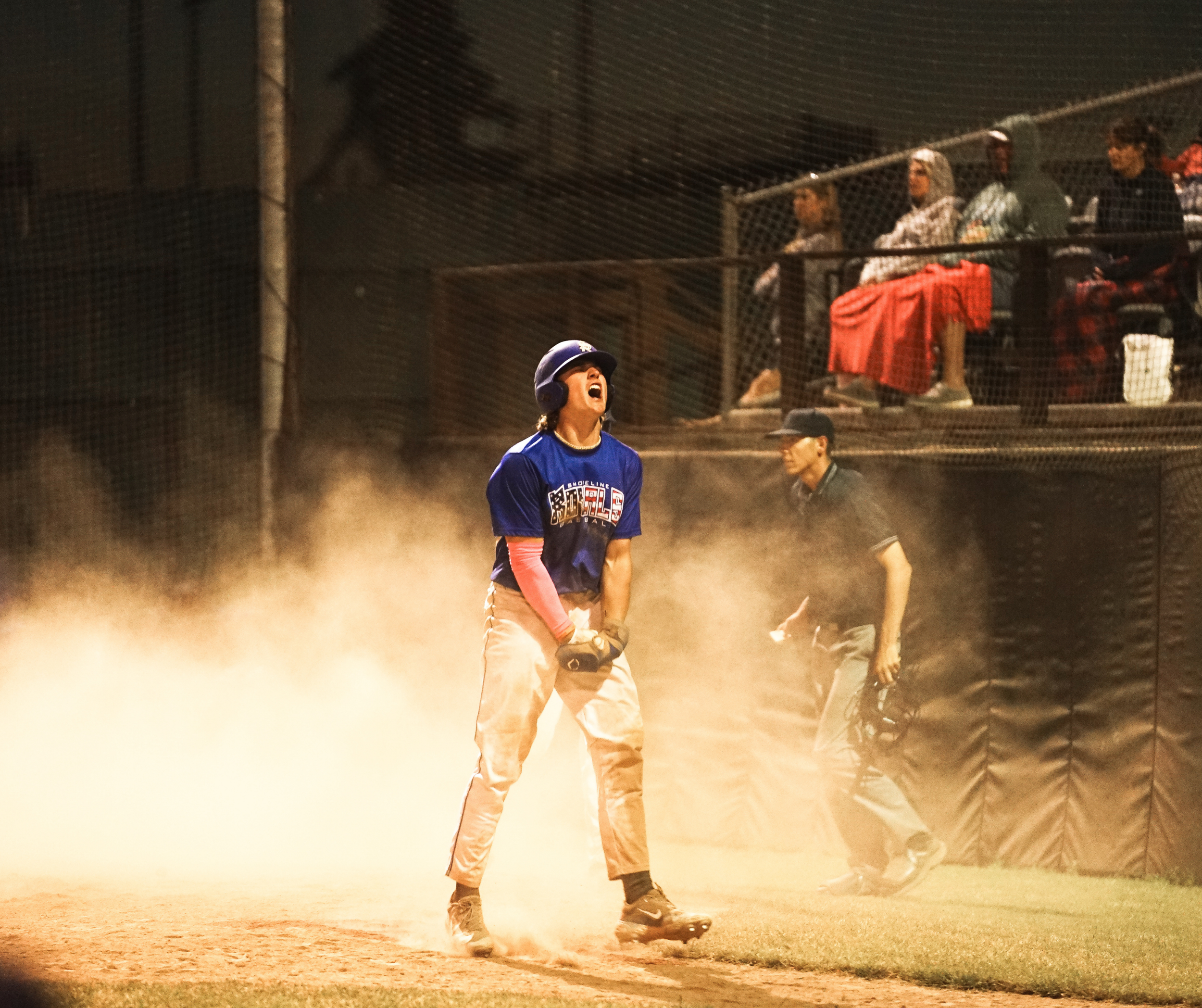 A baseball player in a blue jersey and helmet celebrating on the field at night, with spectators watching from the stands behind a protective fence, illuminated by stadium lights.