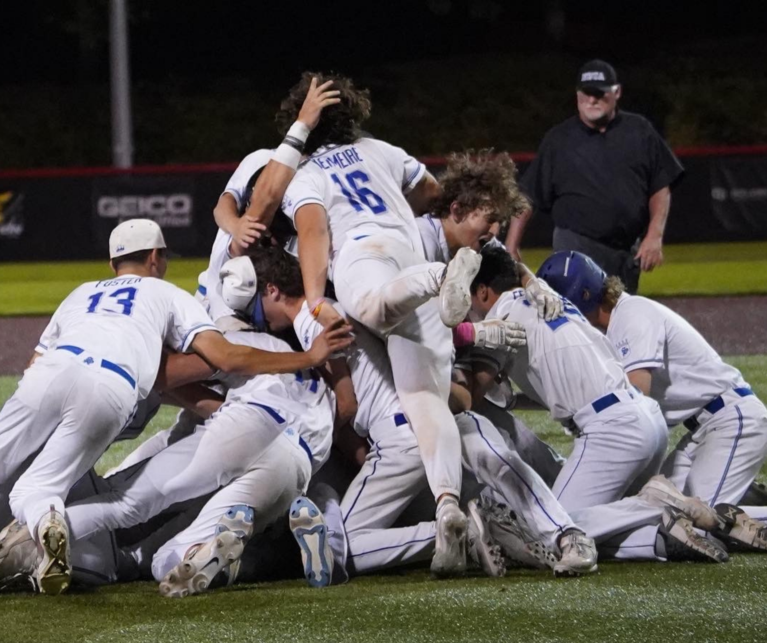 A group of baseball players in white uniforms celebrating on the field, with one player on top and others on the ground, and an umpire standing nearby during a night game.