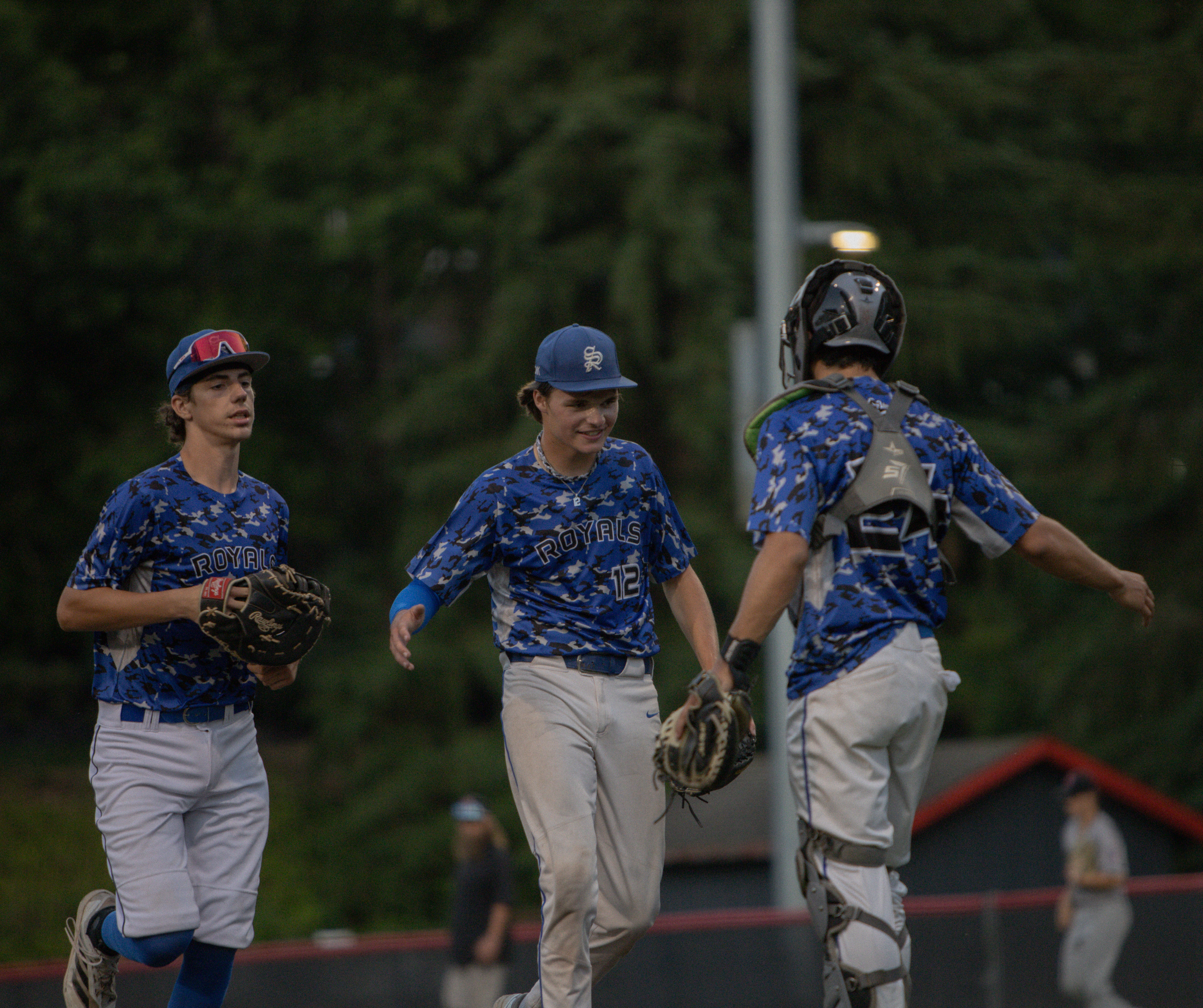 A group of young male baseball players in matching blue camouflage uniforms with 'Royals' written on them, standing outdoors on a grassy field during the evening with trees in the background.